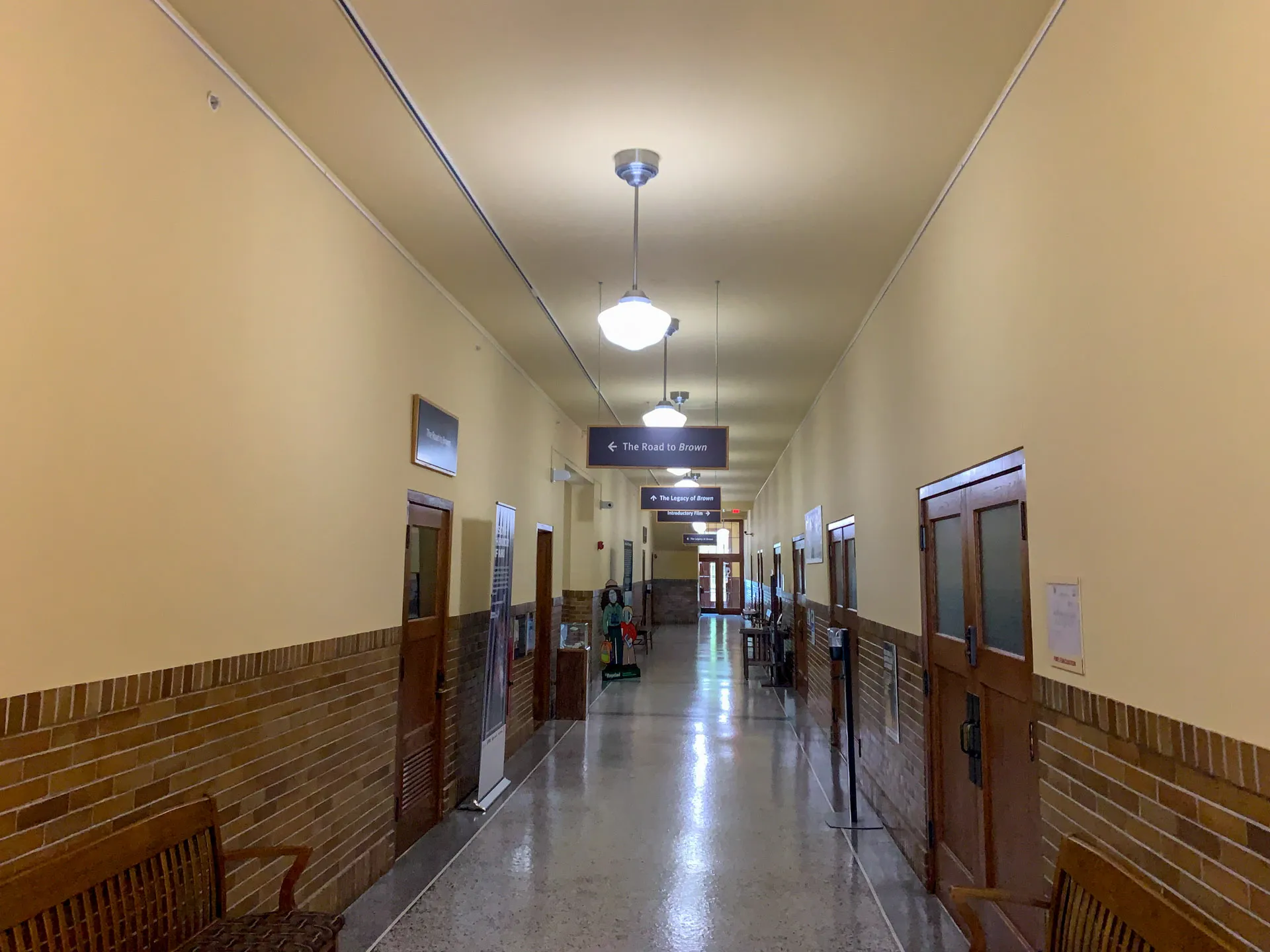 A quiet indoor hallway with cream walls and a shiny tiled floor. Wooden doors with glass panels line the corridor, and a brick accent runs along the lower wall. Overhead, several lights illuminate the space. A sign hangs from the ceiling near the center of the image.