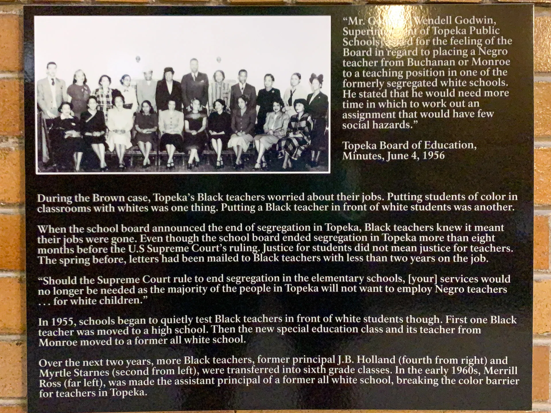 A historical display featuring a brick wall background with an educational plaque. The plaque includes a quote from the Topeka Board of Education dated June 4, 1956, mentioning a Supreme Court case and discussing racial integration in schools. The plaque is surrounded by black and white photos.