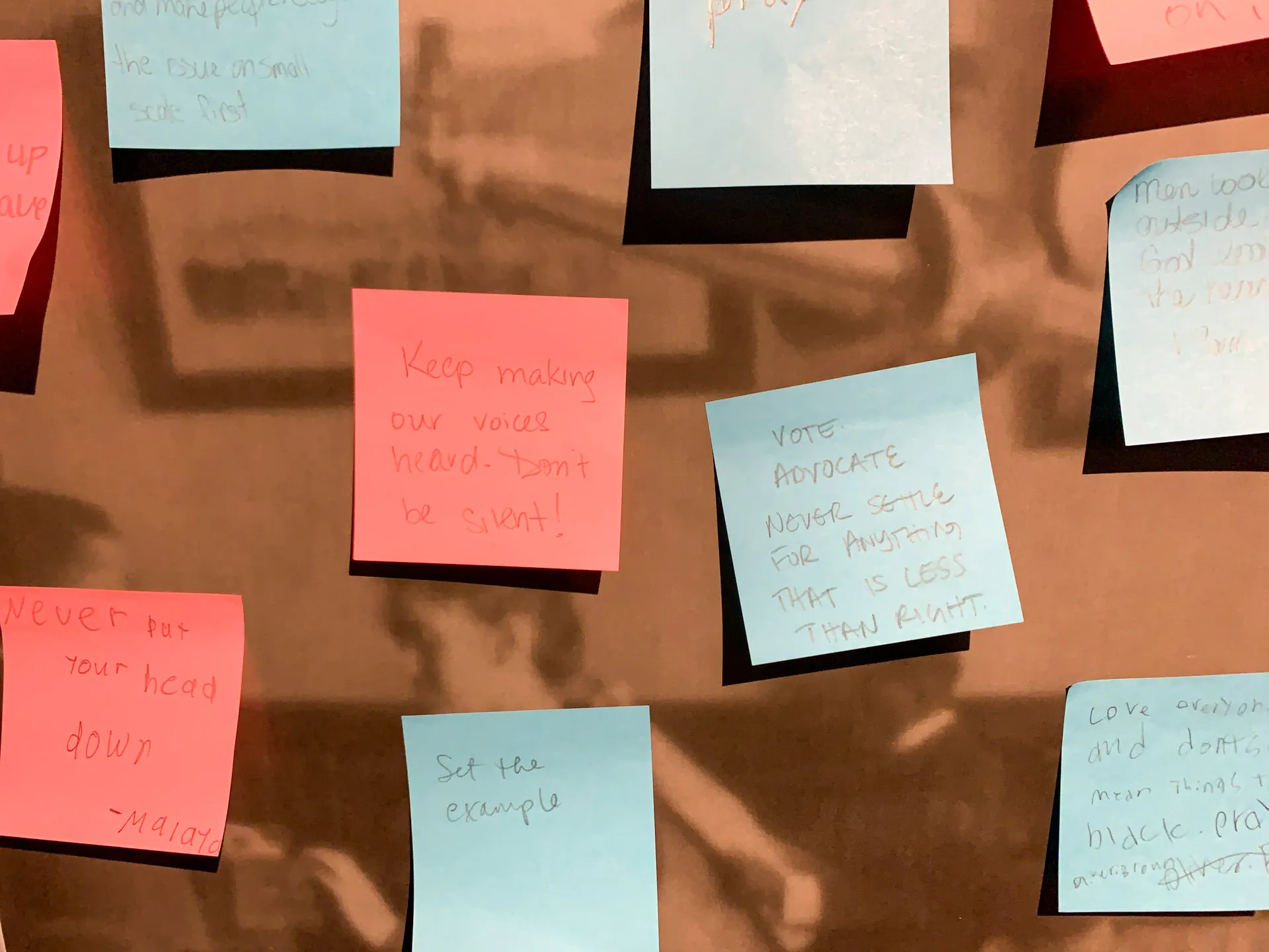 A close-up of a bulletin board covered with various colored sticky notes. The notes include messages like "Keep making our voices heard. Don't be silent!" on a pink note and "Vote, advocate, never settle for anything that is less than right!" on a blue note. Shadows are visible in the background.