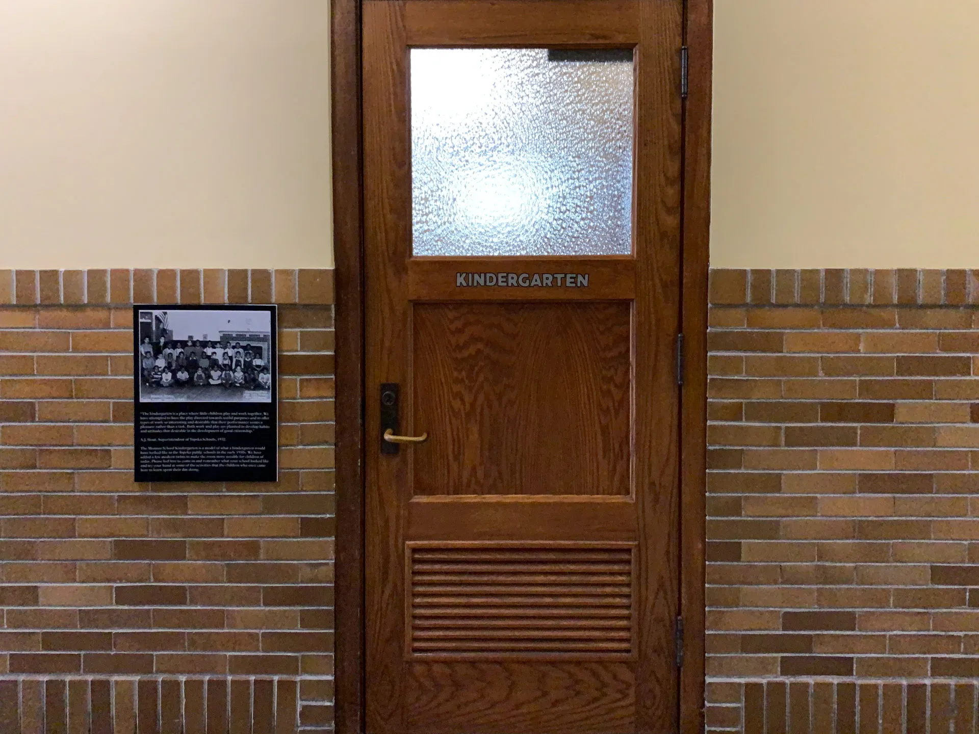 A wooden door labeled "KINDERGARTEN" with a textured glass window is set in a brick wall. To the left, a black plaque with a historic photo and text is mounted on the wall. The lower part of the door features horizontal vents. The wall has dark brown bricks with lighter grout.