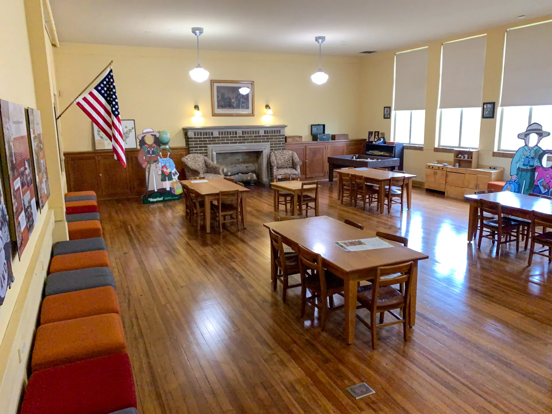 A large, empty classroom with wooden flooring and beige walls features several wooden tables and chairs. An American flag stands in the corner. Colorful murals of children and three illuminated ceiling lights add decor. Windows with blinds line the wall, letting in natural light.