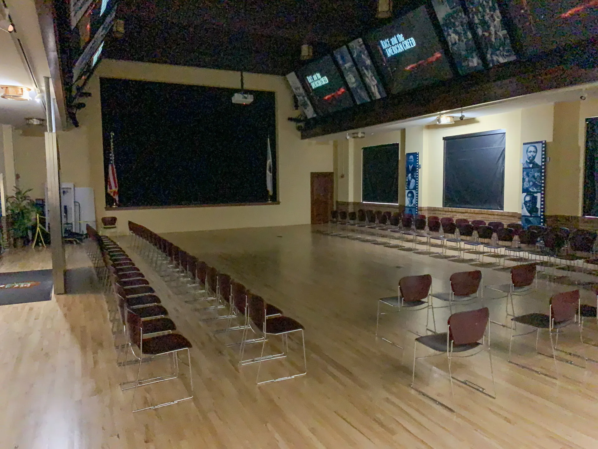 A dimly lit auditorium with rows of empty chairs facing a stage with a large black screen. The wooden floor reflects the muted lighting. Two vertical banners with historical images are visible on the right wall, and an American flag stands near the stage on the left.