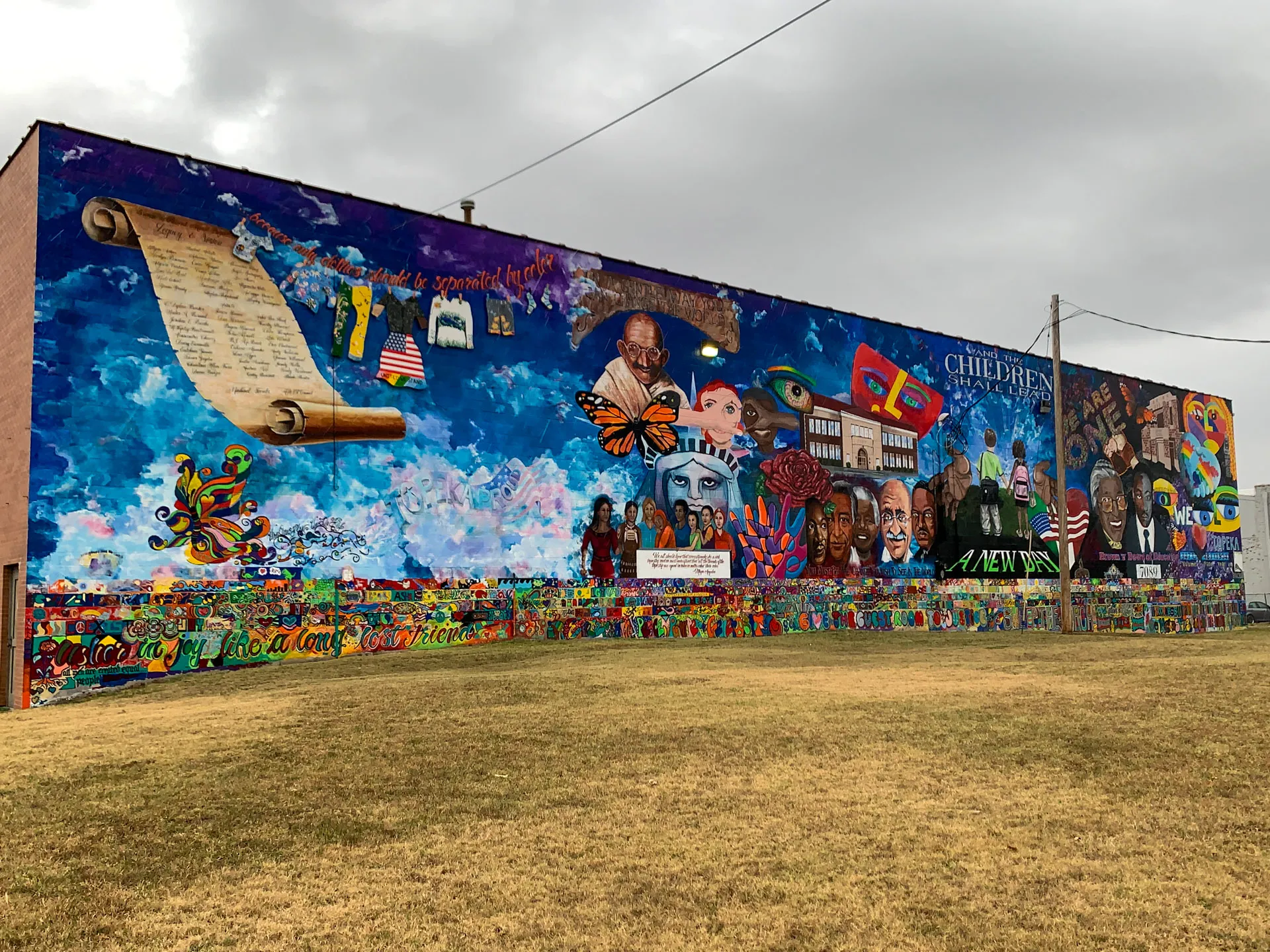 A large, colorful mural on a brick wall features historical figures, a scroll, and vibrant symbols. The sky is depicted with clouds and a butterfly. Diverse faces and scenes convey themes of unity and progress. The grass in the foreground contrasts with the overcast sky above.