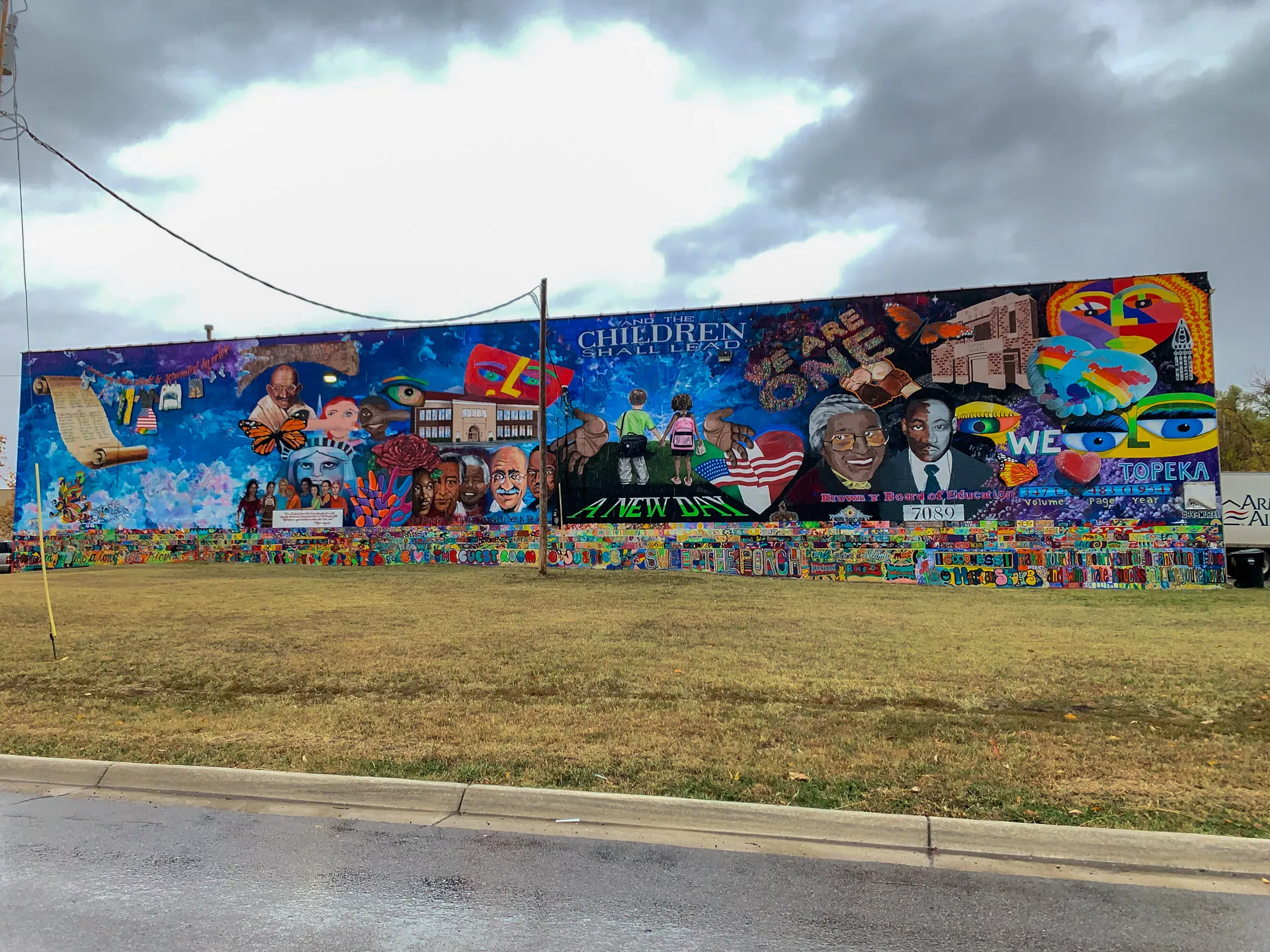 A large outdoor mural on a building facade features colorful imagery including children, diverse faces, musical elements, and symbolic elements like peace signs and butterflies. The mural conveys themes of unity, peace, and community engagement. The foreground shows a grassy area and street.