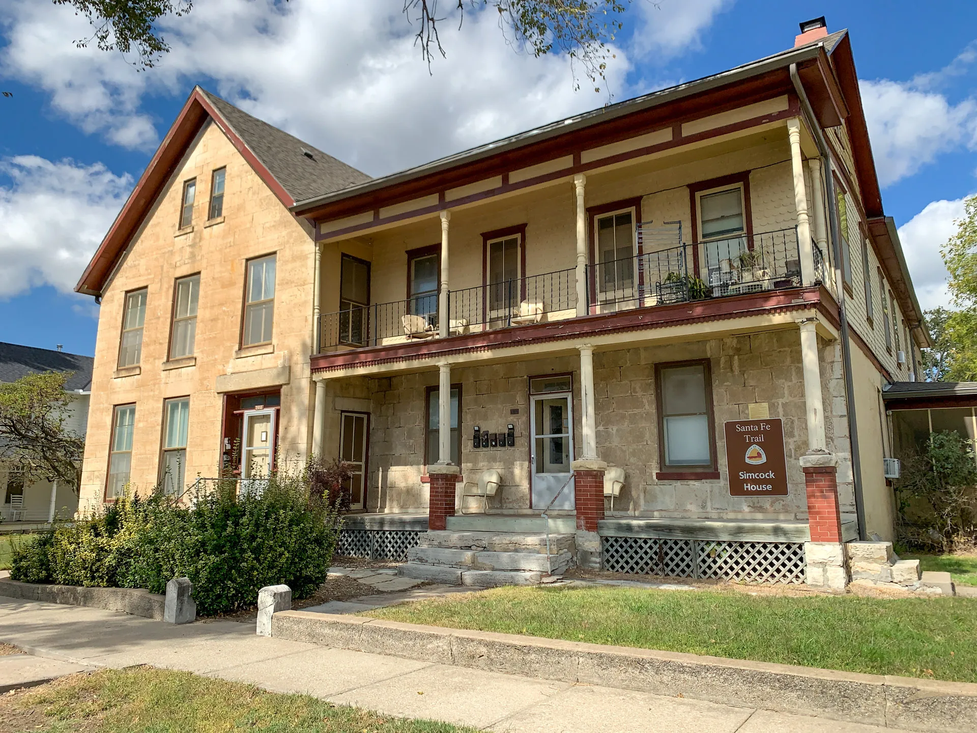 Historic limestone building with two stories, red trim, and a covered balcony. A sign reads "Simeon & Jane Stewart House." The front yard has shrubs, concrete steps, and a pathway. The sky is partly cloudy, enhancing the classic facade.