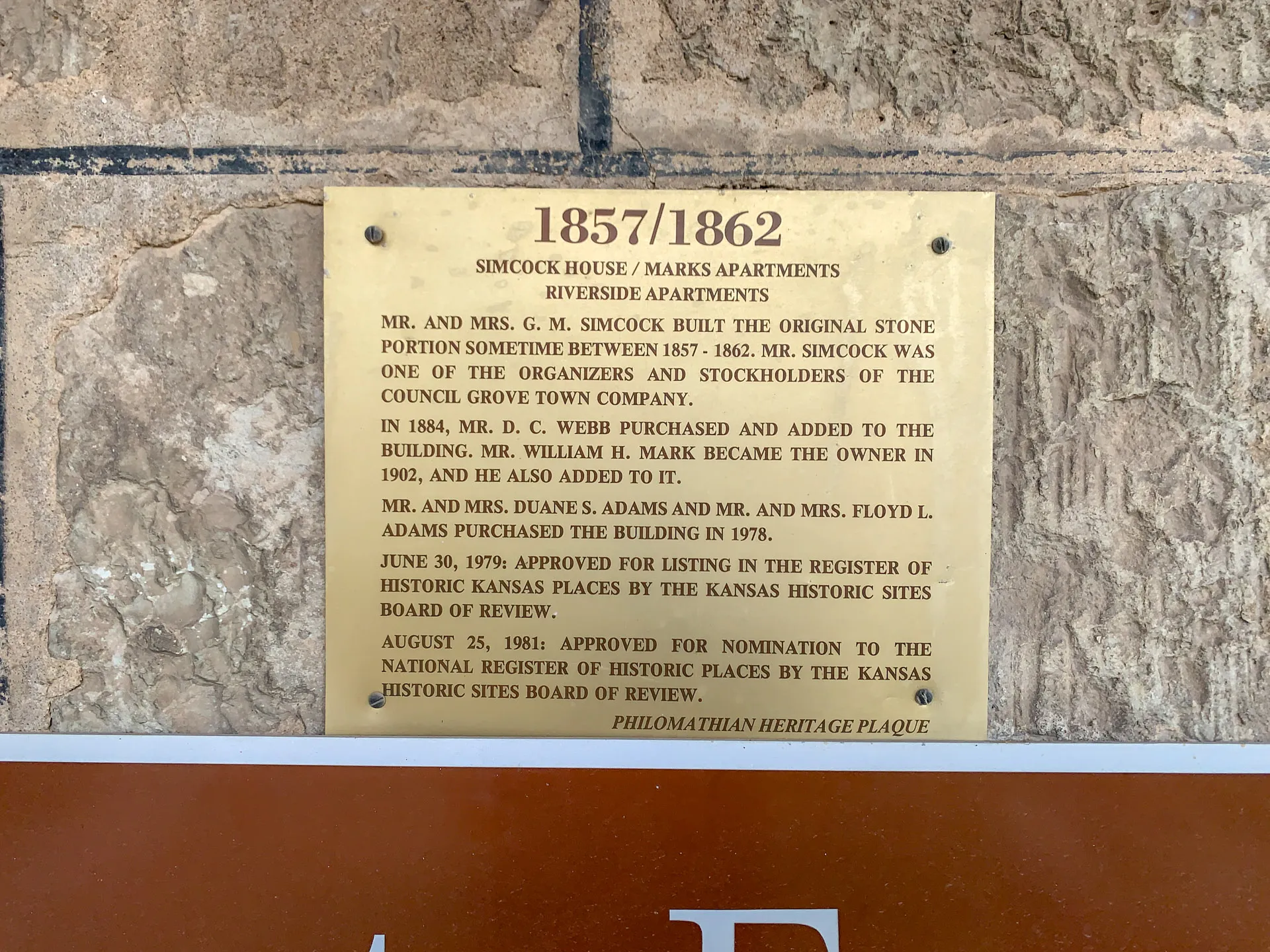 A rectangular brass plaque mounted on a stone wall. It details the history of the Simcock House/Marrs Apartments, mentioning owners and renovations from 1857 to 1979. The plaque notes nominations for historic registers in 1979 and 1981 by the Kansas Historical Society.