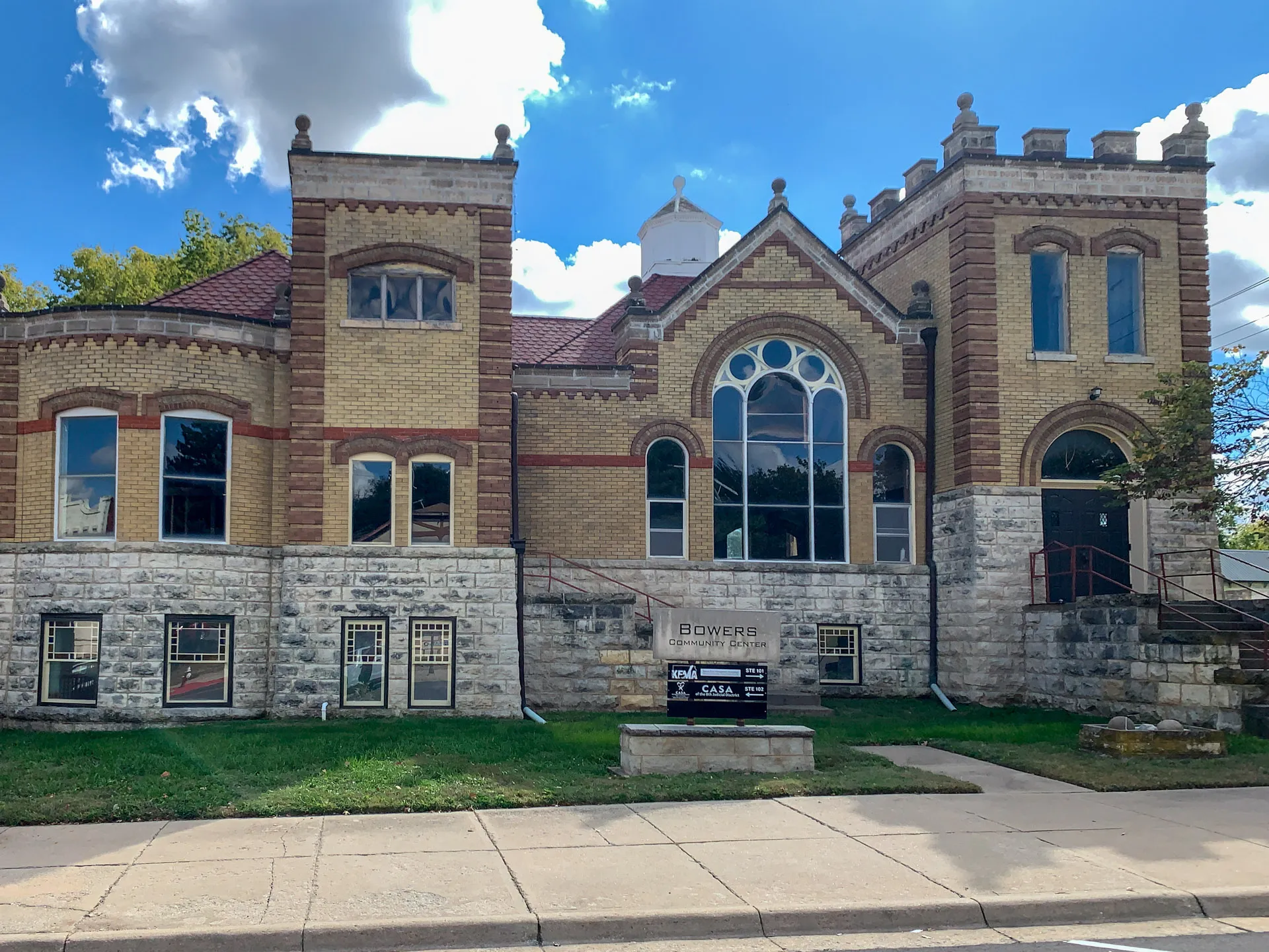 A historic, castle-like building with tan brick and stone foundation, featuring arched windows and a crenelated roofline. A brick pattern adds visual interest. A sign in front reads "Bowers." The building is set against a partly cloudy sky, with a sidewalk and street in the foreground.