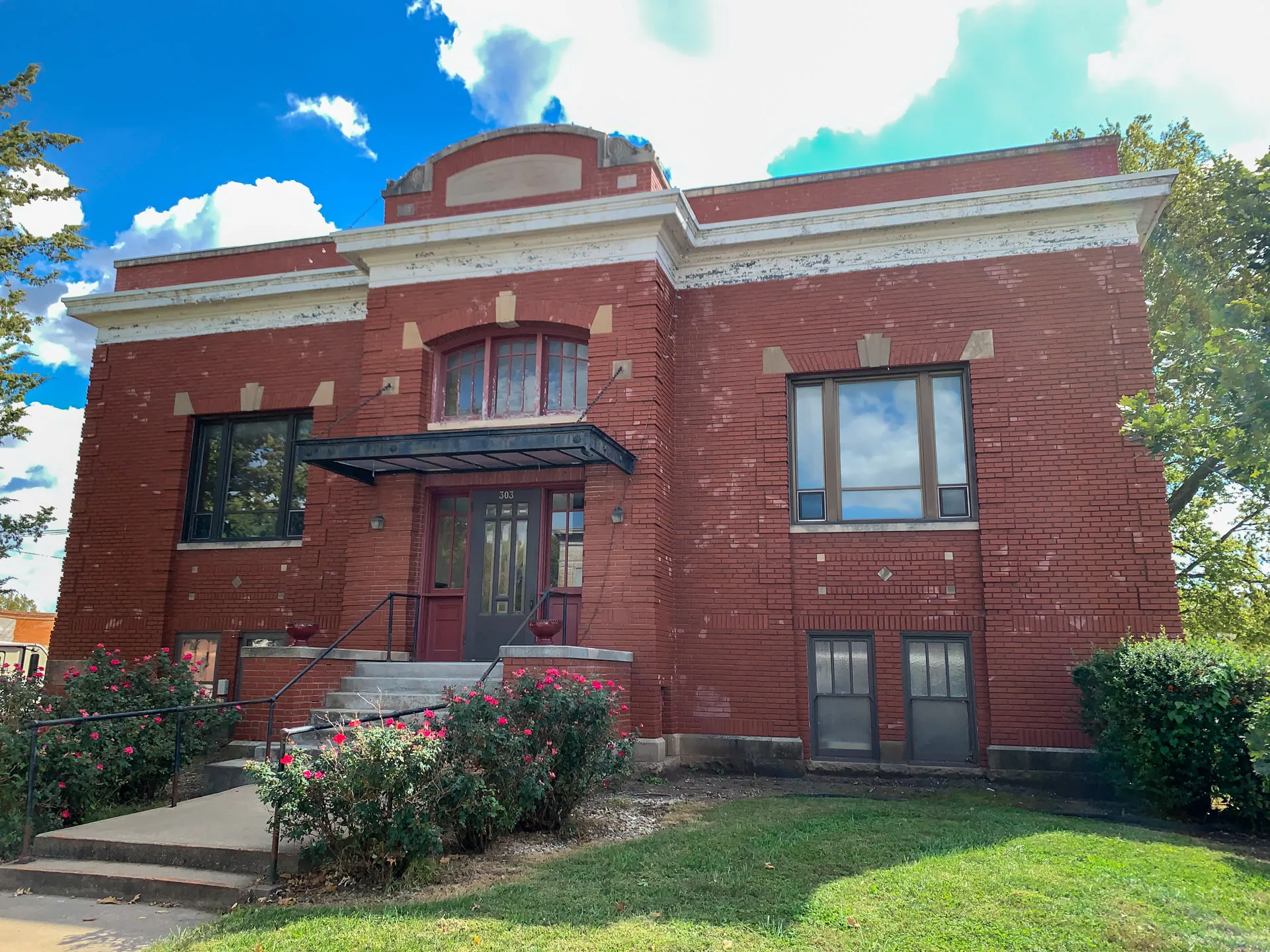 A historic red brick building with a central entrance, featuring a dark awning and stairs leading up. Two trees frame the structure, with neatly trimmed bushes and vibrant pink flowers in front. The sky is bright blue with fluffy white clouds, adding to the scene's charm.