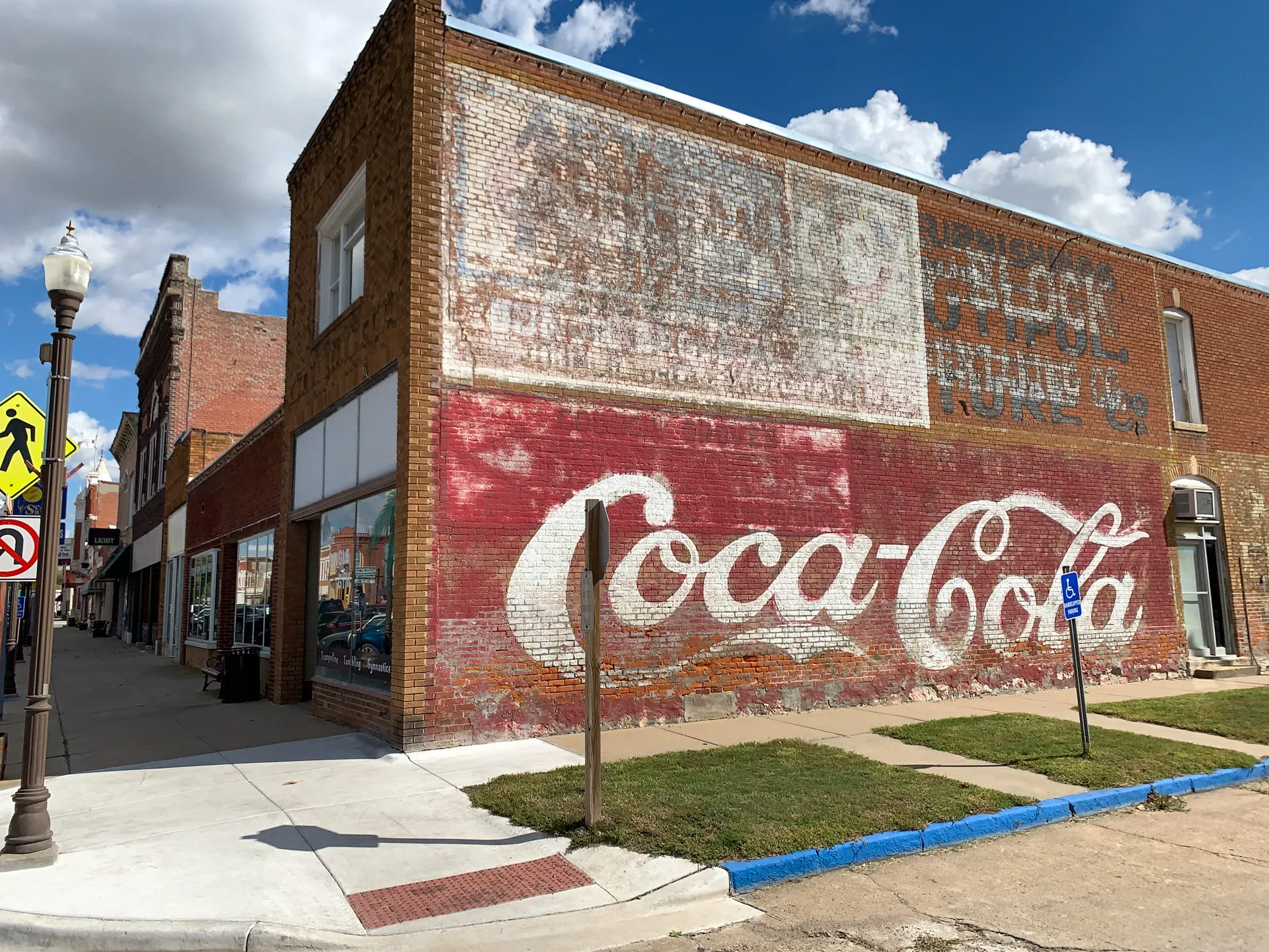 A vintage brick building features a large, faded Coca-Cola mural. The mural covers the side wall in red and white. The street corner includes a sidewalk with reflective store windows and a lamppost. A bright blue sky with fluffy clouds is visible above.
