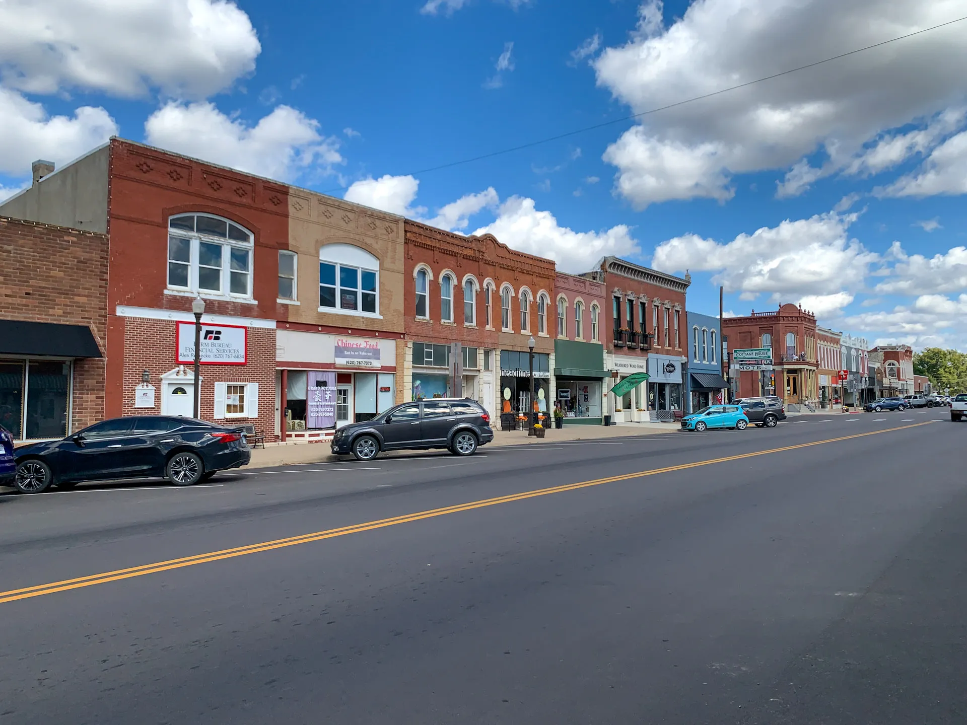 A vibrant small-town street lined with historic brick buildings under a blue sky dotted with fluffy clouds. Cars are parked alongside the road, and people are strolling along the sidewalk, creating a lively yet relaxed atmosphere. The architecture features ornate facades and varied designs.