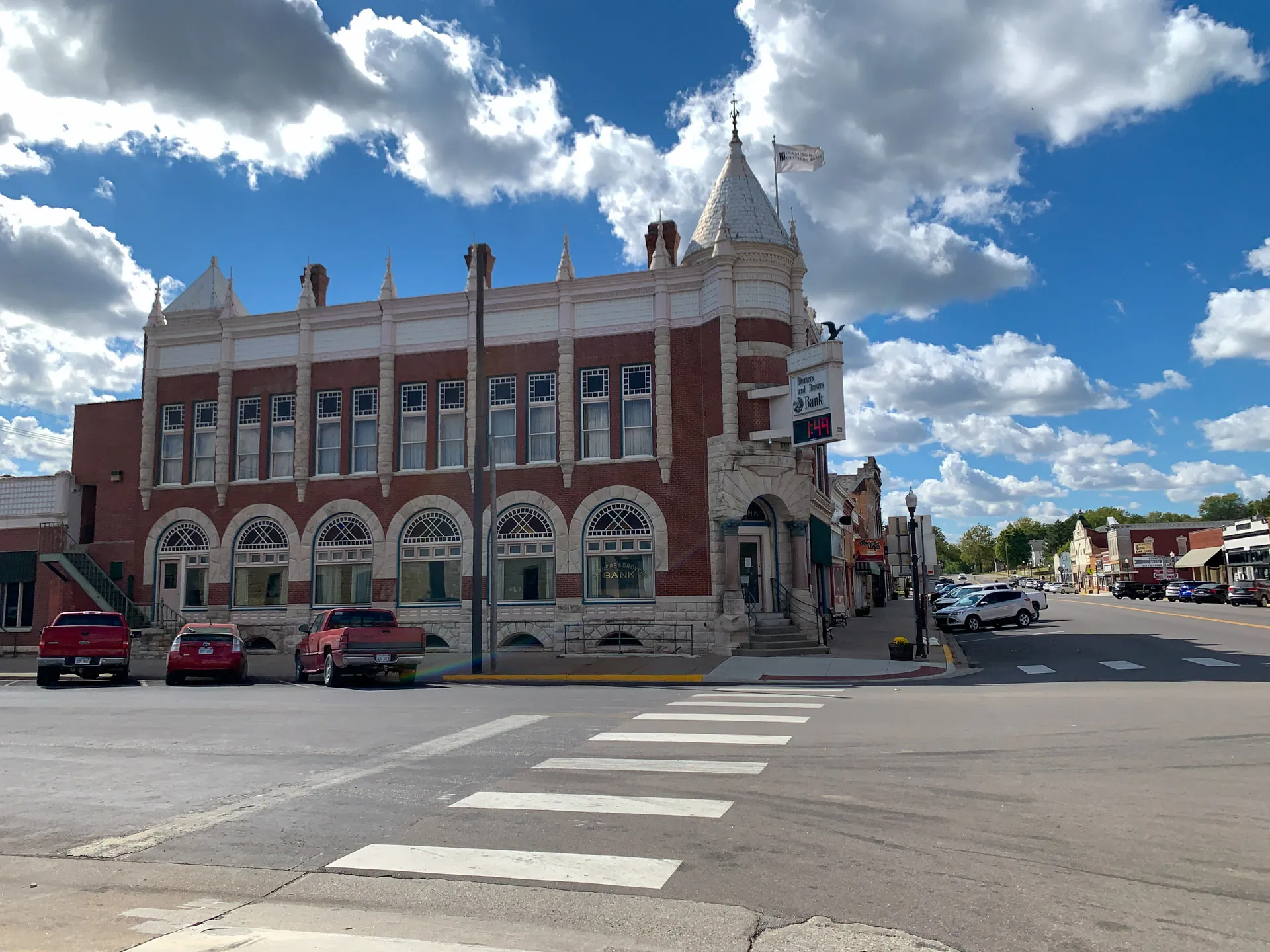 A historic, ornate brick building with arched windows and a turret stands on a street corner under a blue sky with fluffy clouds. Several parked cars line the street, which features a white crosswalk in the foreground. The building houses a jewelry store, as indicated by a visible sign.