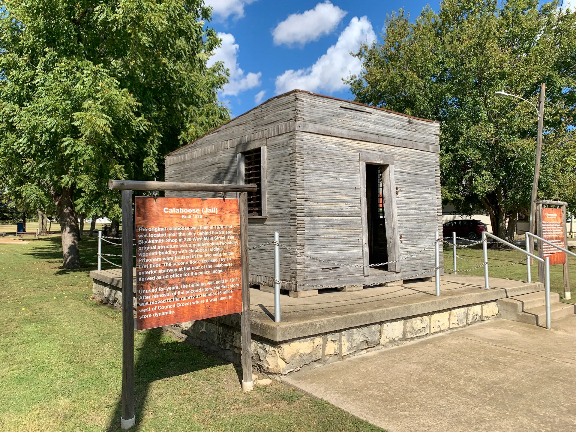 A small, weathered wooden jail building, known as Calaboose, is set on a stone foundation under a bright blue sky. A brown informational sign is displayed in the foreground, and the area is surrounded by trees and grass. A path leads to the entrance, which is secured by a chain barrier.