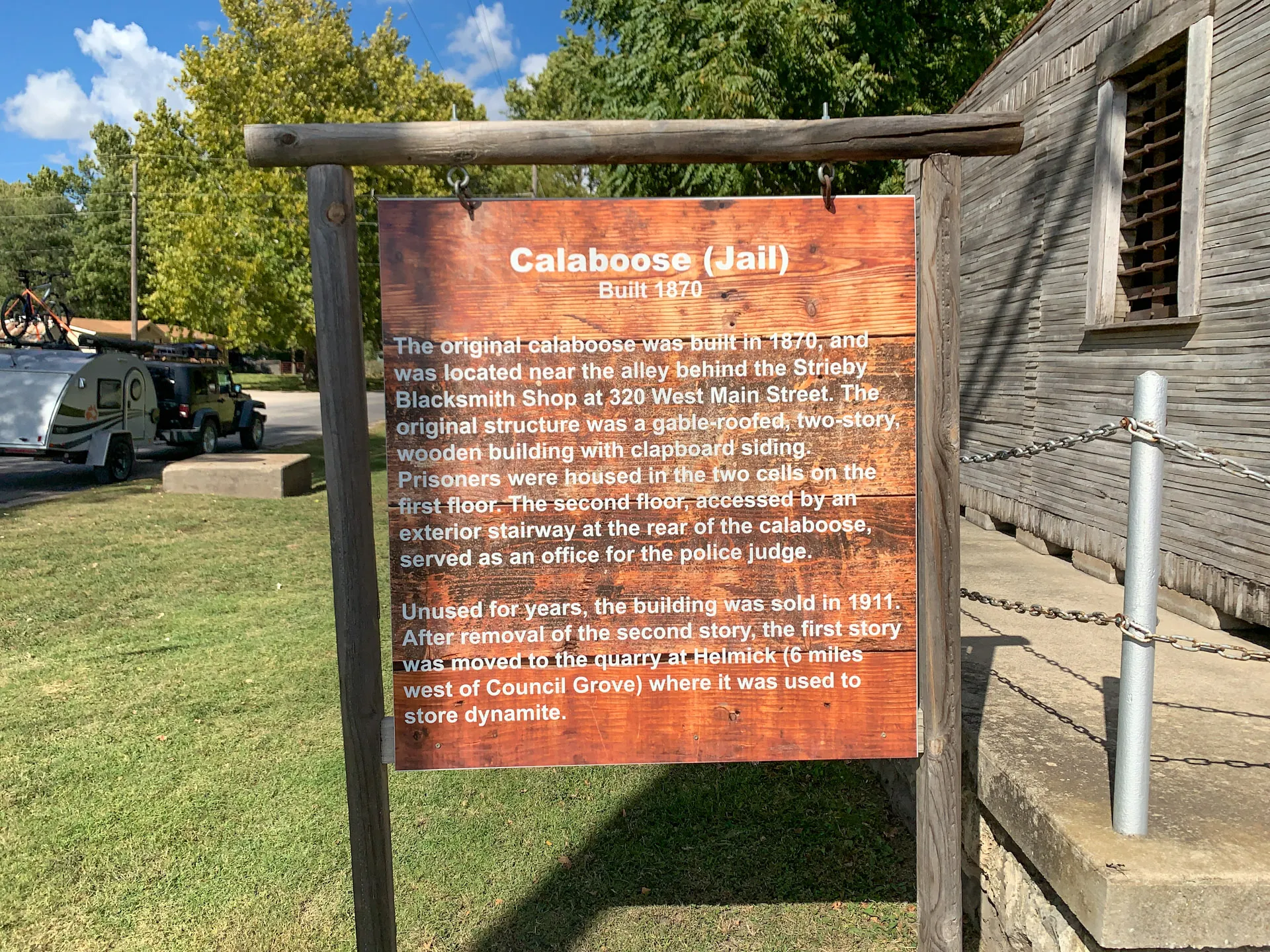 A wooden sign titled "Calaboose (Jail) Built 1870" stands against a sunny backdrop, detailing the history and features of an old jail, including its original structure and later use as a storeroom for dynamite. A pickup truck and trees are visible in the background.