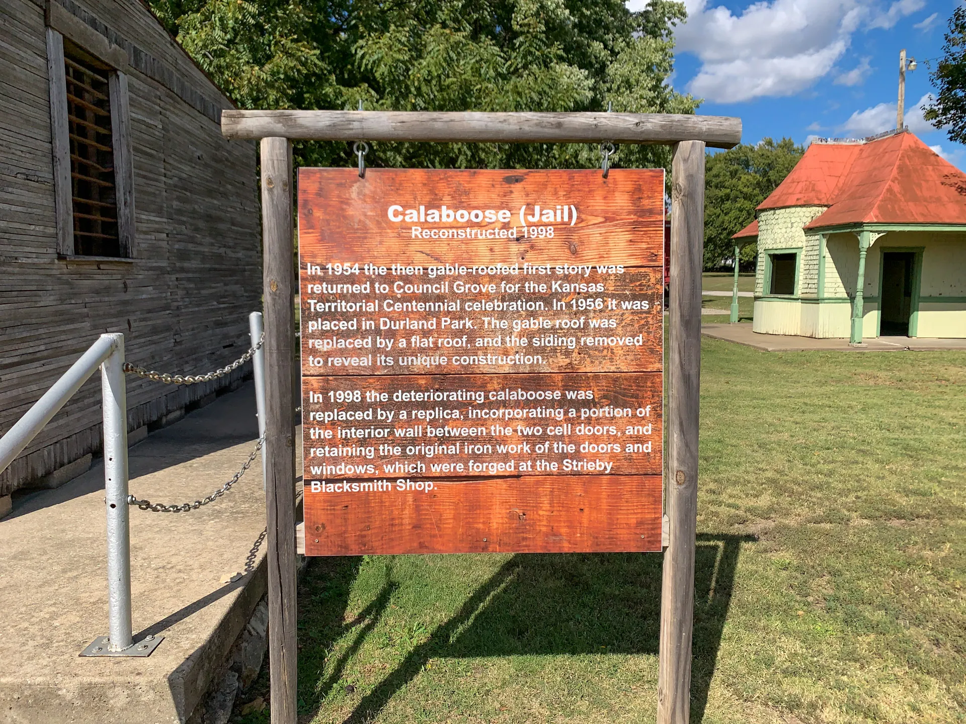 A wooden sign in front of a wooden building reads: "Calaboose (Jail) Reconstructed 1998." It details the history of the jail, mentioning its use in the 1954 Kansas Territorial Centennial and reconstruction in 1998. A park with a green lawn and a red-roofed structure is visible in the background.