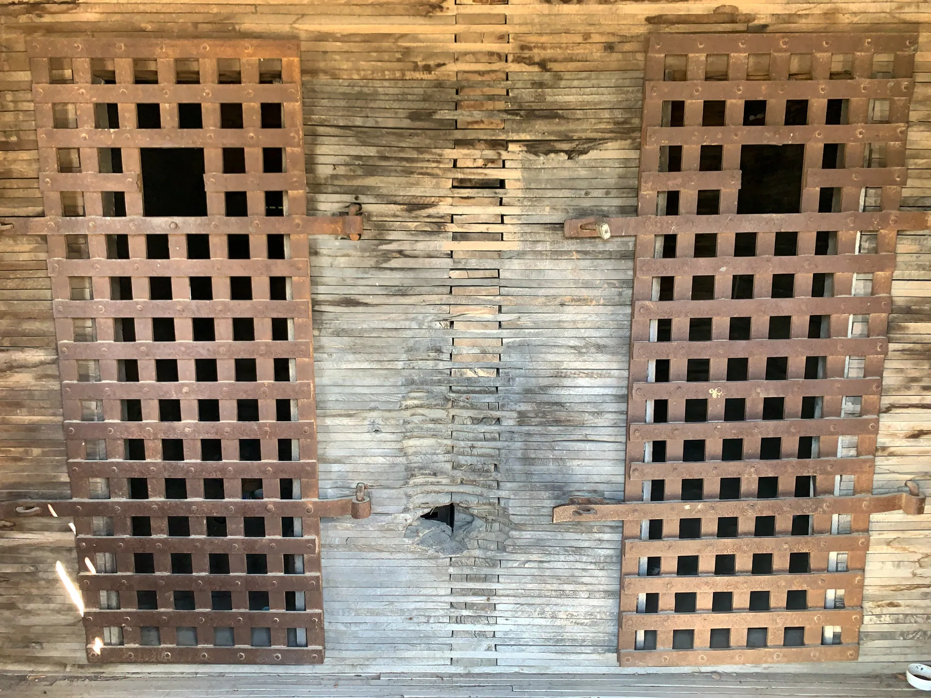Rustic wooden wall with two large, rusty metal grates, each with square openings. The grates are mounted side by side, secured with metal latches. A small hole is present between them at the bottom of the wall, revealing wear and age. The slats are unevenly spaced and discolored.