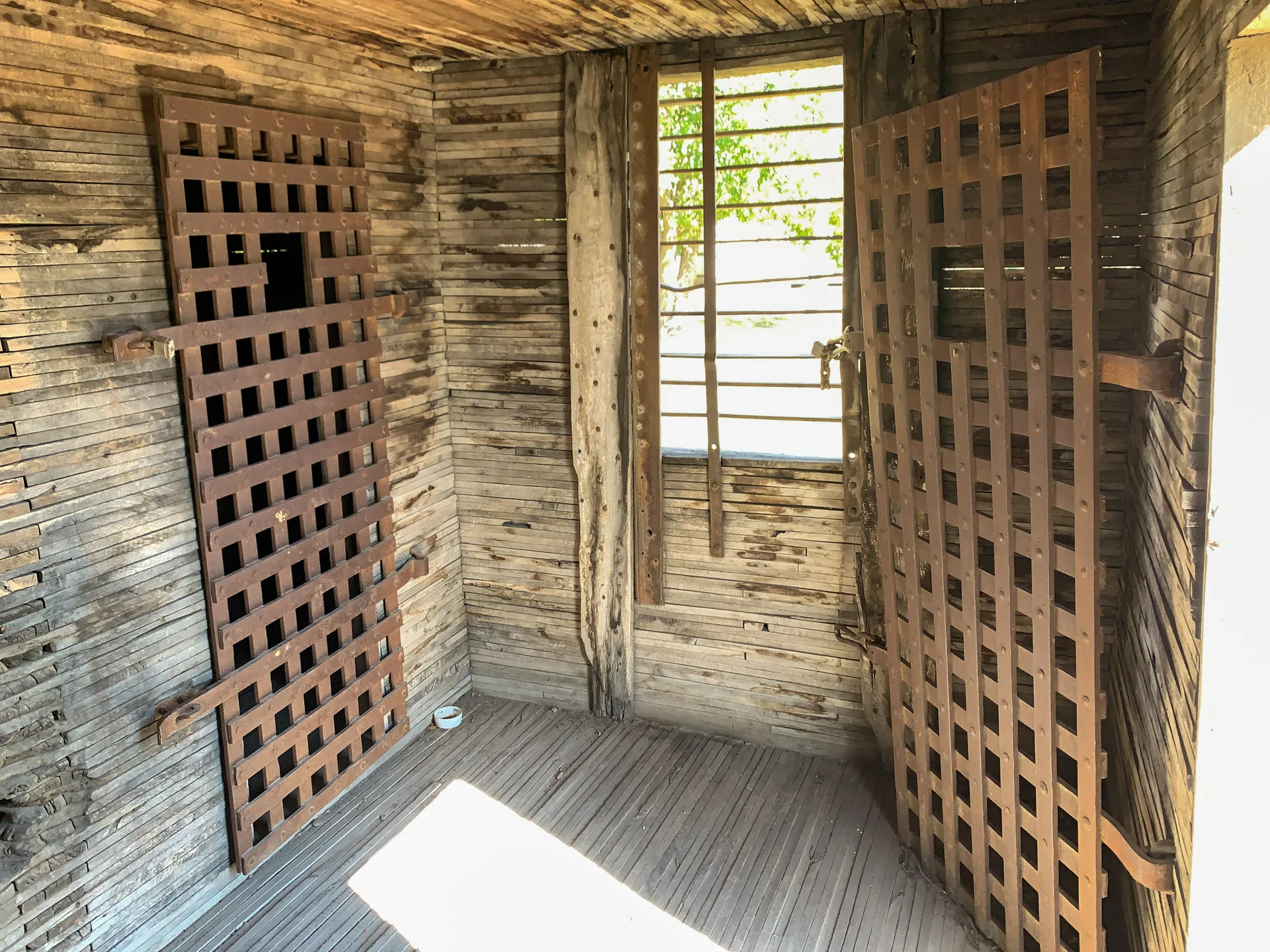 A small wooden jail cell with two barred doors is shown. The walls and floor are made of weathered wood. One door is ajar, and there is a barred window with sunlight streaming in, casting shadows. A small metal tin is on the floor near the open door.