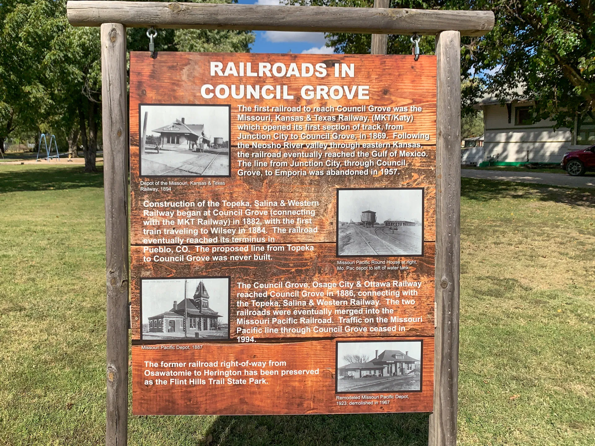 A wooden sign in a park titled "Railroads in Council Grove," with historical text and black-and-white photos depicting old trains and stations. It describes the development and importance of the Missouri-Kansas-Texas and Council Grove railroads. Trees and grass are visible in the background.