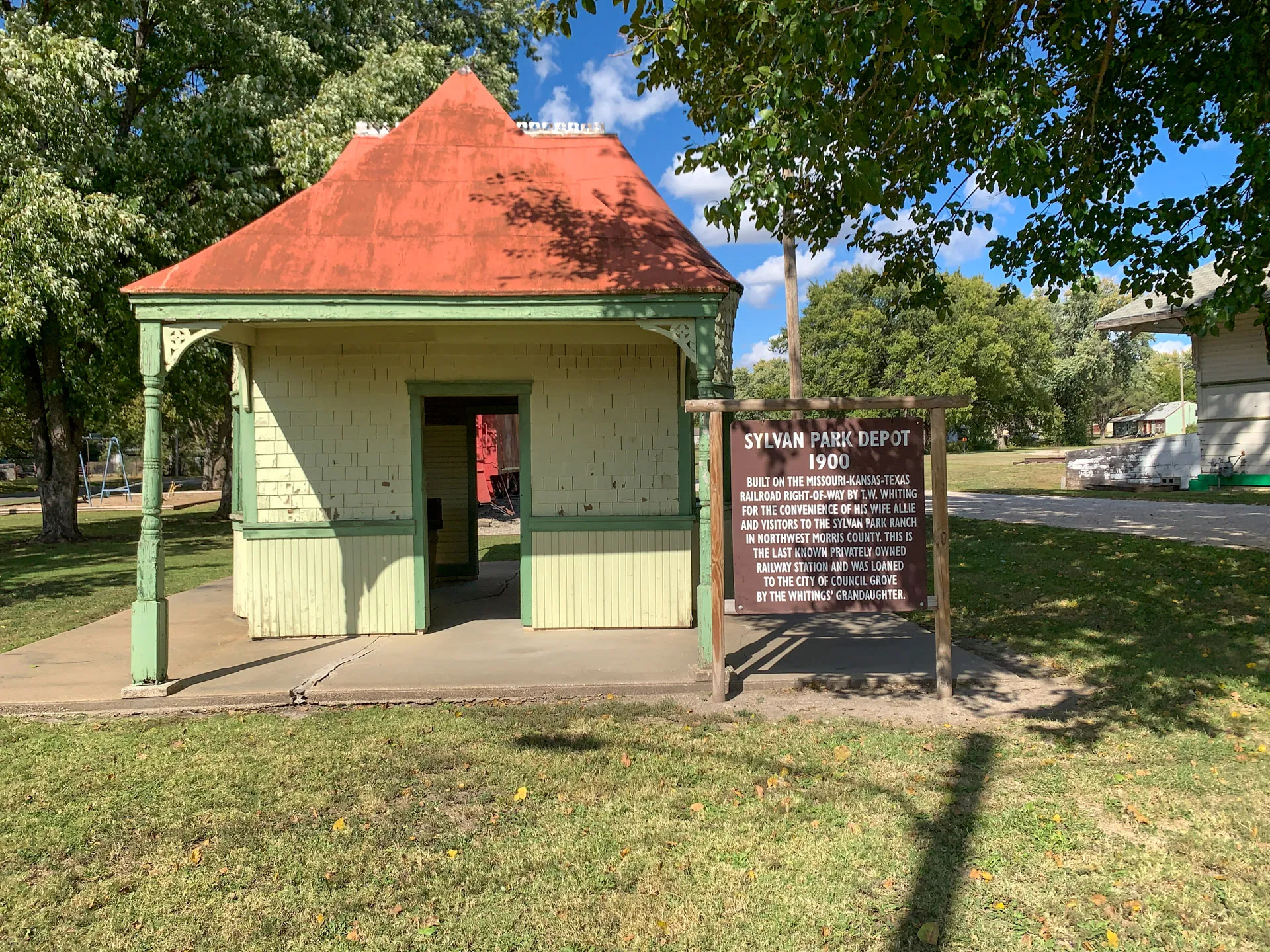 A small historical building with a red roof and green walls stands in a park. A wooden sign next to it reads "Sylvan Park Depot 1900," with additional text detailing its history. The area is grassy with trees and a clear blue sky in the background.