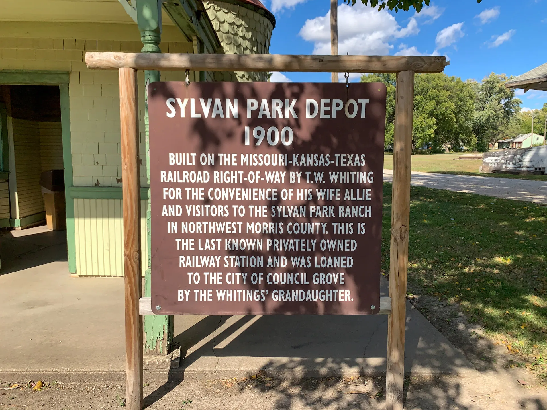 A wooden sign reads: "Sylvan Park Depot 1900. Built on the Missouri-Kansas-Texas railroad right-of-way by T.W. Whiting for the convenience of his wife Allie and visitors to the Sylvan Park Ranch in northwest Morris County. This is the last known privately owned railway station and was loaned to the City of Council Grove by the Whitings' granddaughter." It stands outside a building under a bright blue sky with trees and grass in the background.