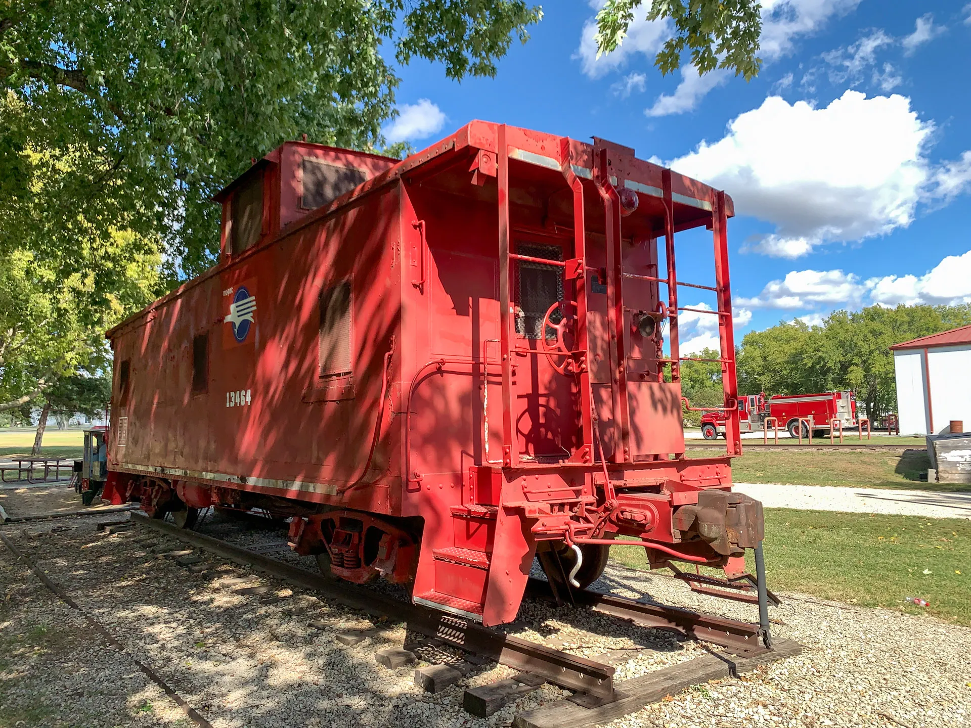 A bright red caboose is situated on short railway tracks in a park-like setting. The sky is clear with a few clouds, and trees provide shade. Nearby, a red building and some grass are visible. The caboose features a small cupola on top, with ladders on the ends and the number 12484.