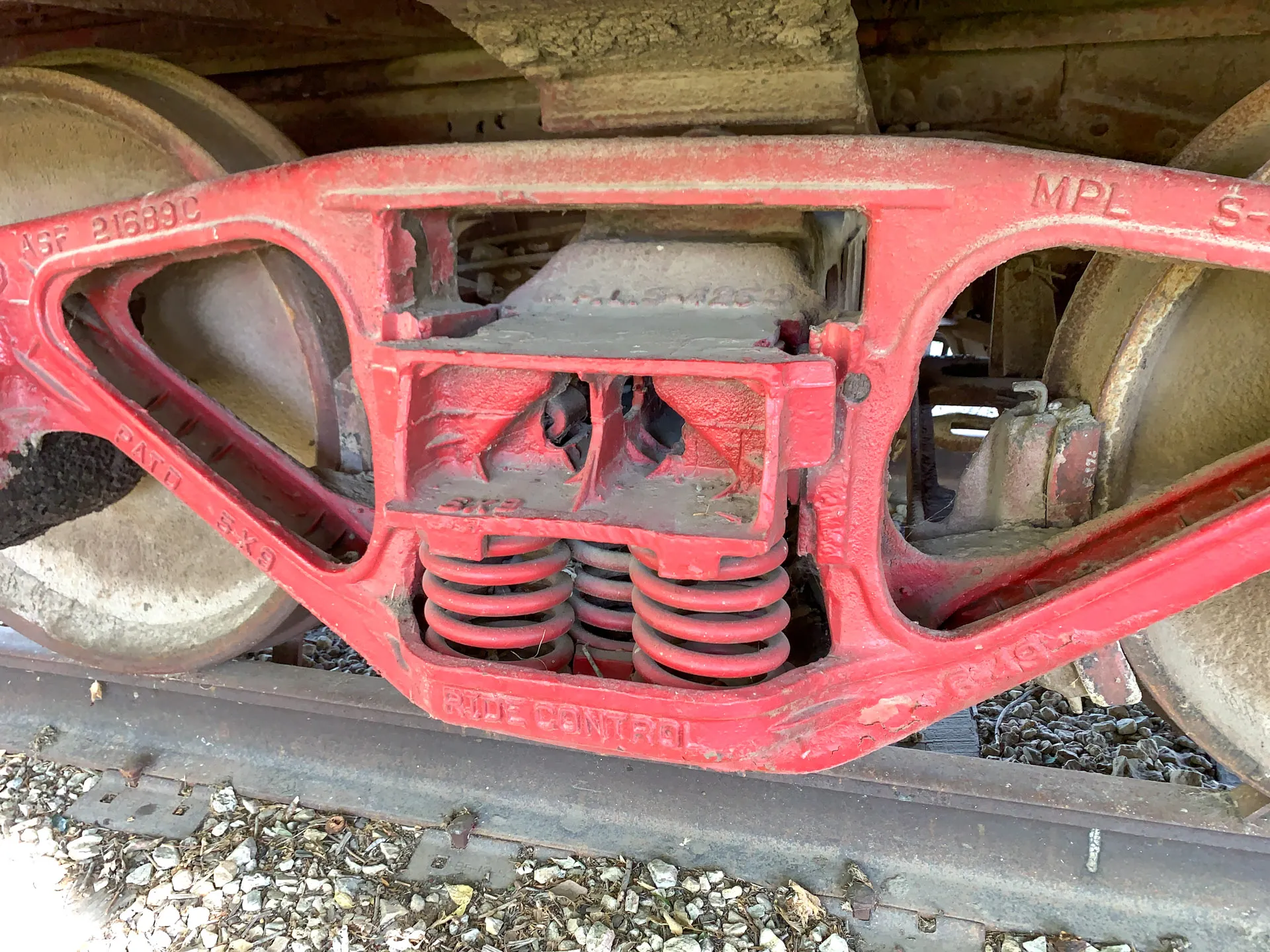 Close-up of a red train wheel and suspension assembly. The metal structure includes a pair of coil springs, with visible wear and grime. The words "Ride Control" and other numbers are embossed on the metal. Gravel and train tracks are seen below.