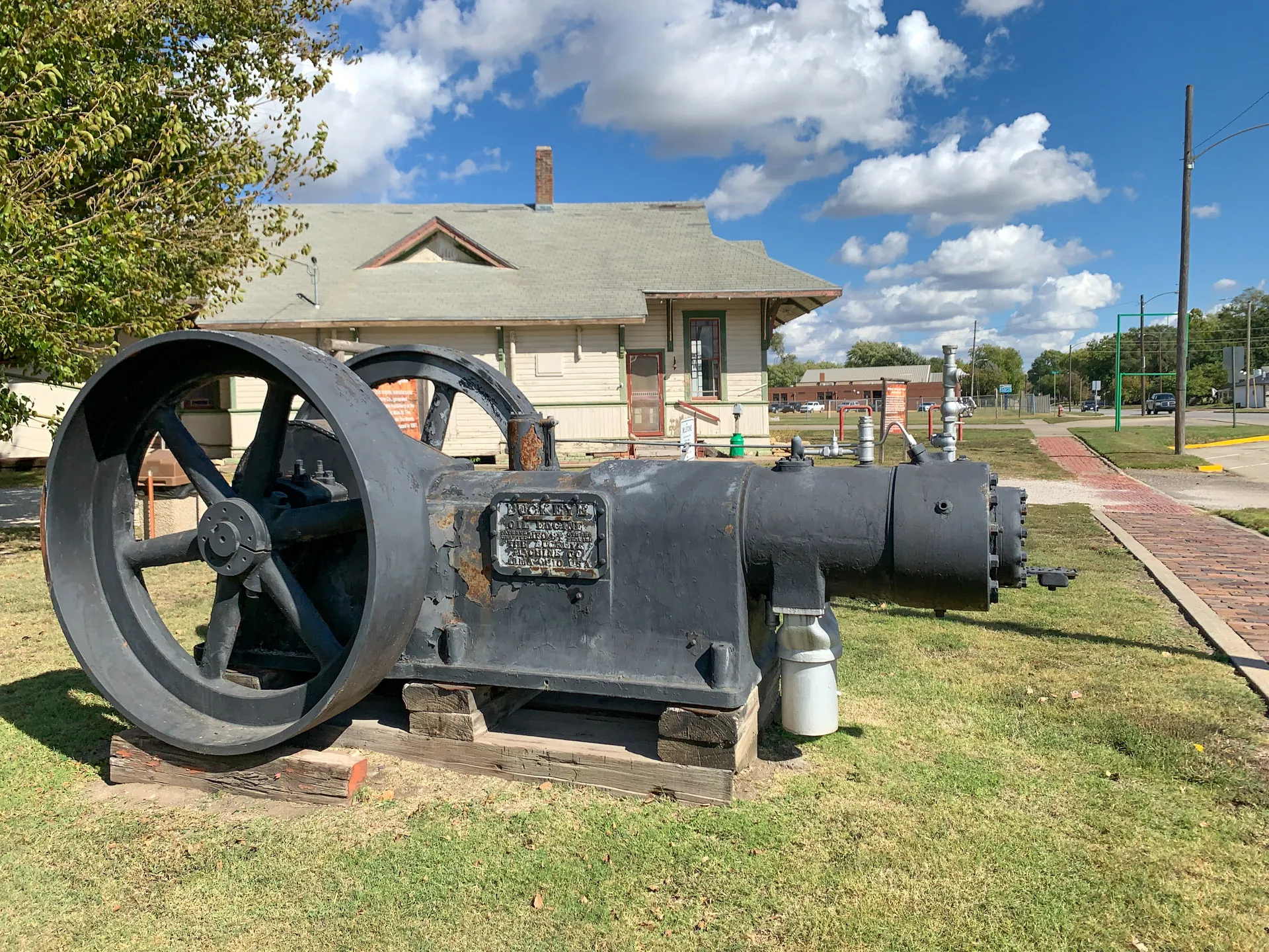 A large, vintage industrial engine sits on wooden blocks outdoors, surrounded by grass. A historic building is in the background under a blue sky with scattered clouds. The engine is metallic with a prominent flywheel. A small white container is placed next to it.