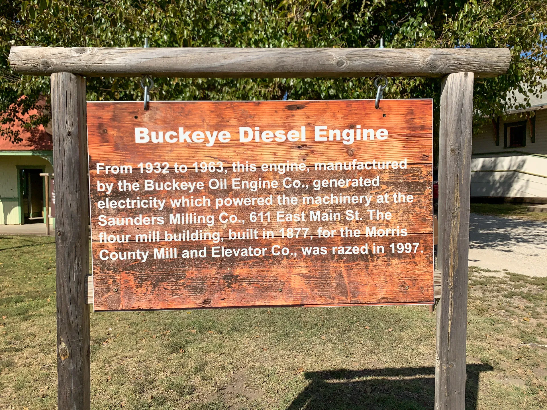 A wooden sign under a tree displays white text. It reads: "Buckeye Diesel Engine: From 1932 to 1963, this engine, manufactured by the Buckeye Oil Engine Co., generated electricity which powered the machinery at the Saunders Milling Co., 611 East Main St. The four mill building, built in 1877, for the Morris County Mill and Elevator Co., was razed in 1997." The sign is part of an outdoor display.
