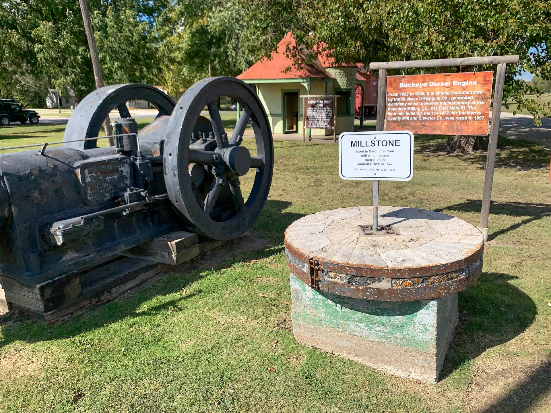 An outdoor display features an old Buckeye diesel engine on the left and a millstone on the right. The engine is black and weathered, with a large wheel. A sign behind it provides historical information. Trees and a small building are visible in the background under clear skies.