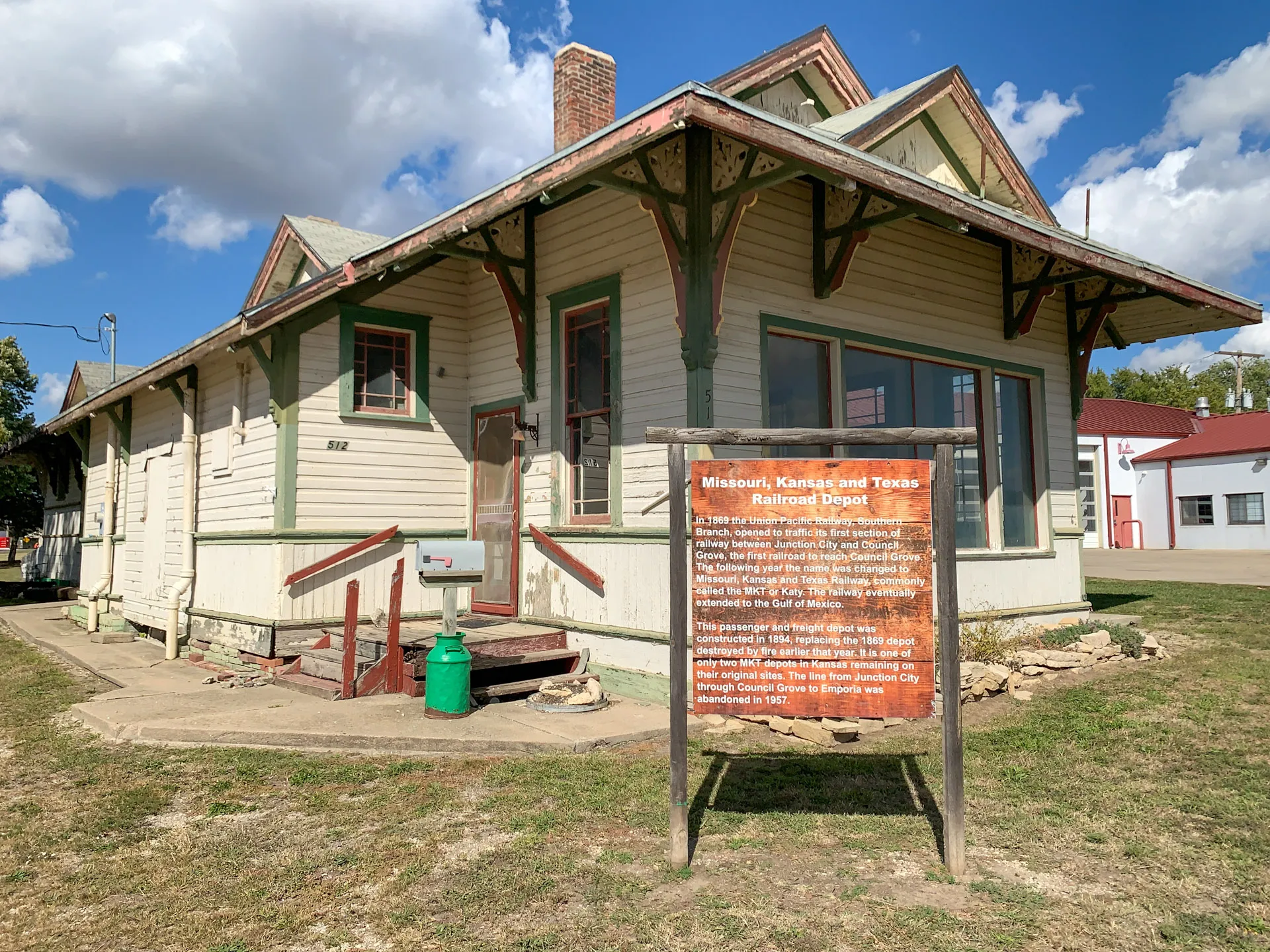 Old wooden railroad depot with a gabled roof, beige siding, and green trim. A historical marker in front provides information about the Missouri, Kansas, and Texas Railroad Depot. The sky is clear with scattered clouds and there's a small red building in the background.
