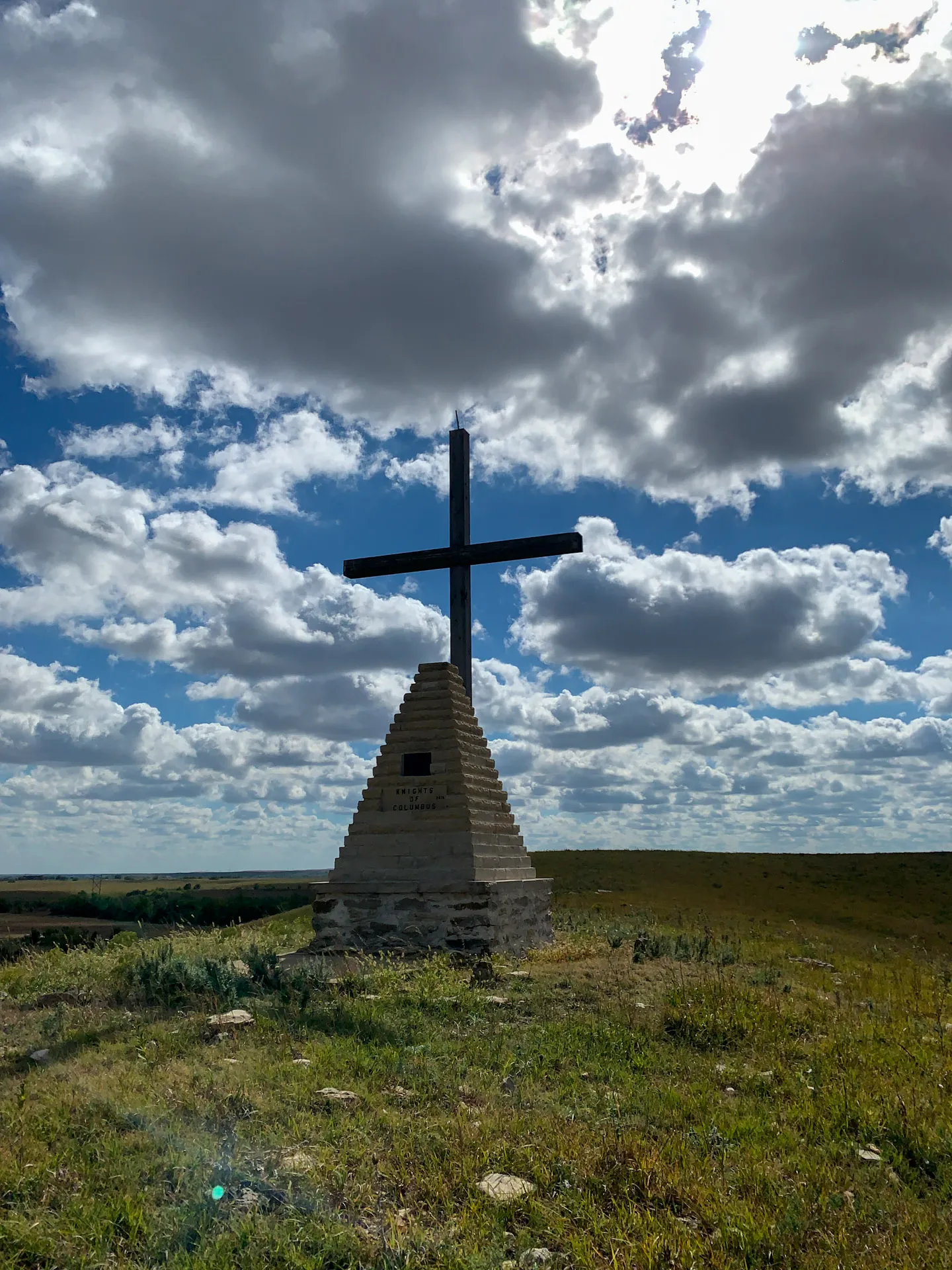 A large stone cross atop a layered, pyramid-like base stands on a grassy hill under a partly cloudy sky. The sun illuminates the scene, casting shadows on the cross and grass. The horizon is visible in the distance, adding depth to the landscape.