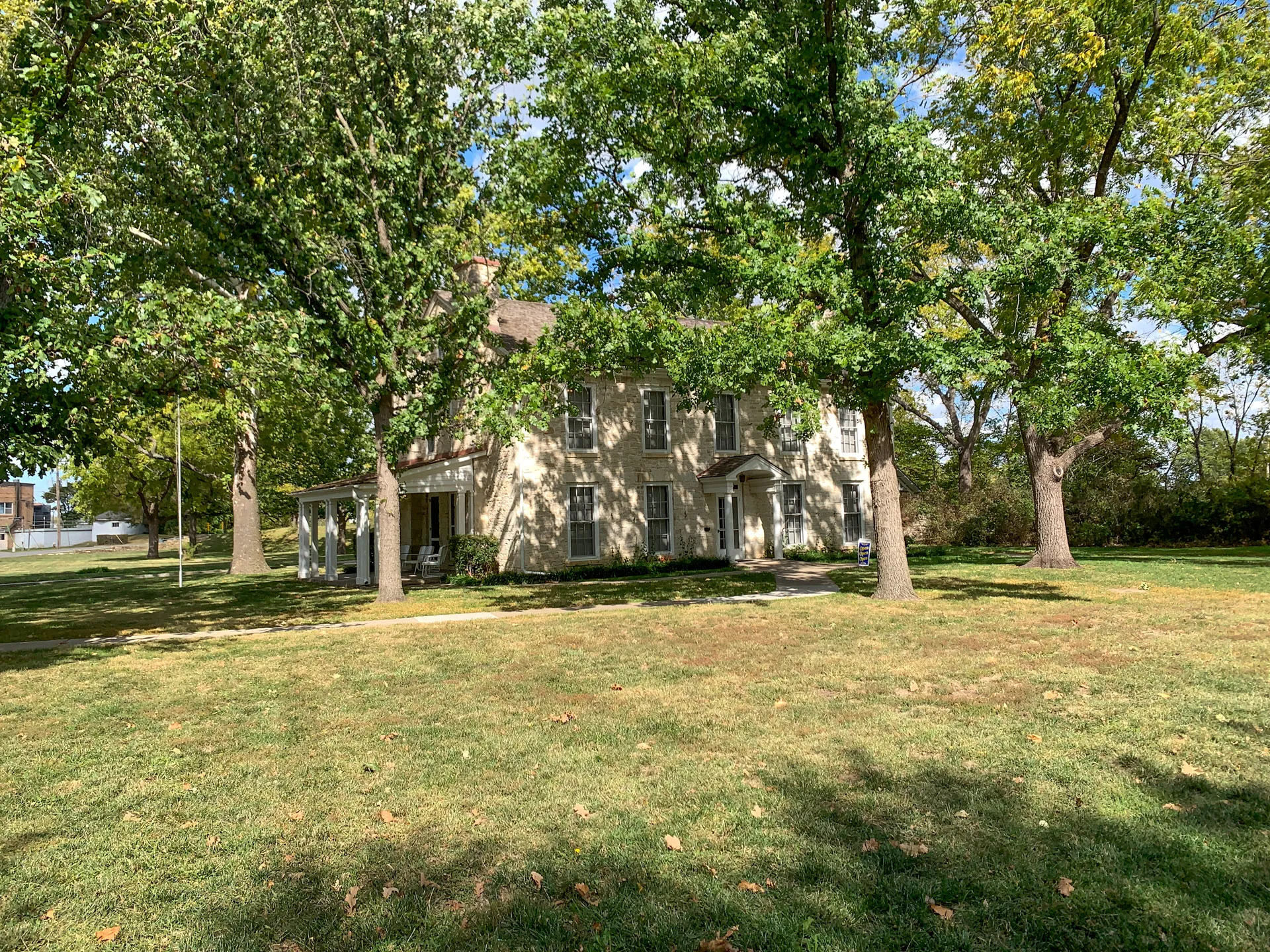 A two-story beige house with white trim is partially obscured by large trees in a grassy area. The trees are lush with green leaves, casting gentle shadows on the lawn and house. A clear blue sky is visible in the background, providing a serene and sunny atmosphere.