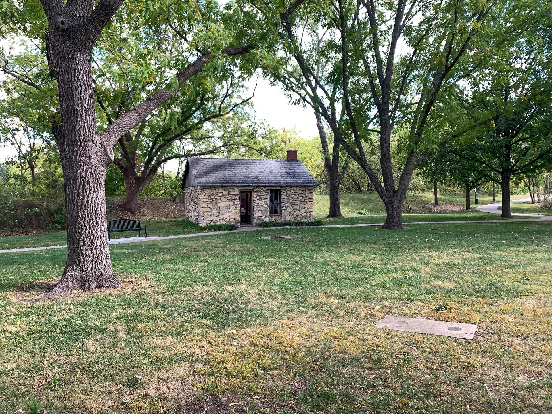 A small stone building with a gray roof is nestled among green trees in a park. A pathway leads to its wooden door. There's a bench on the left side of the path. The surrounding grass is green with patches of brown. The scene is peaceful and bathed in natural light.