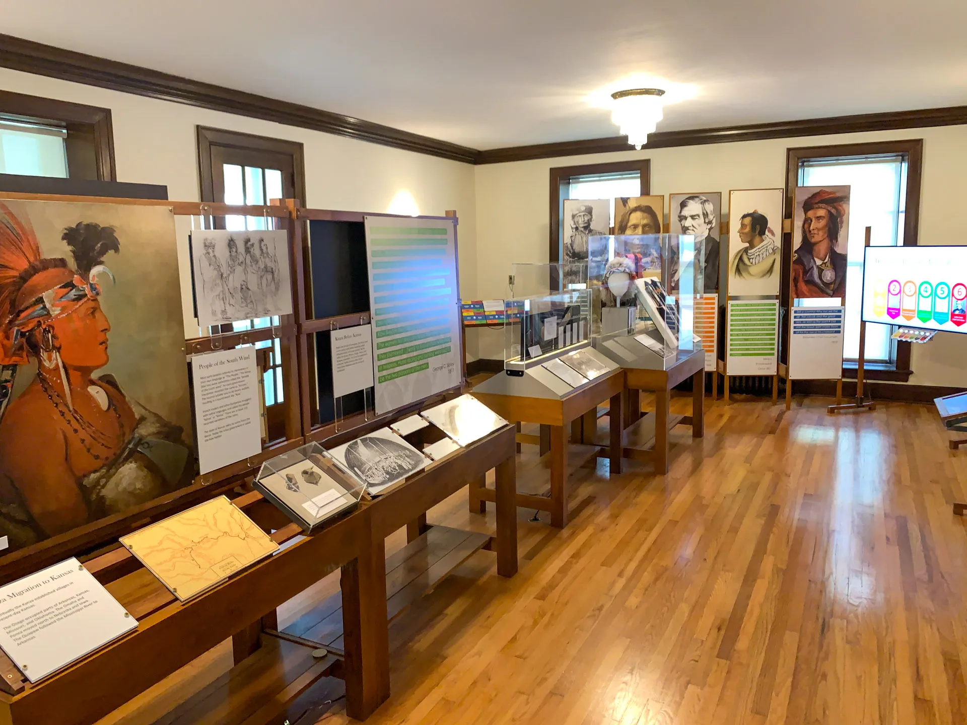 A museum exhibit room with wooden floors and a variety of displays. Art panels and information boards are arranged neatly, showcasing historical illustrations and artifacts. A digital screen displays colorful graphs. The walls are lined with large portraits and windows.