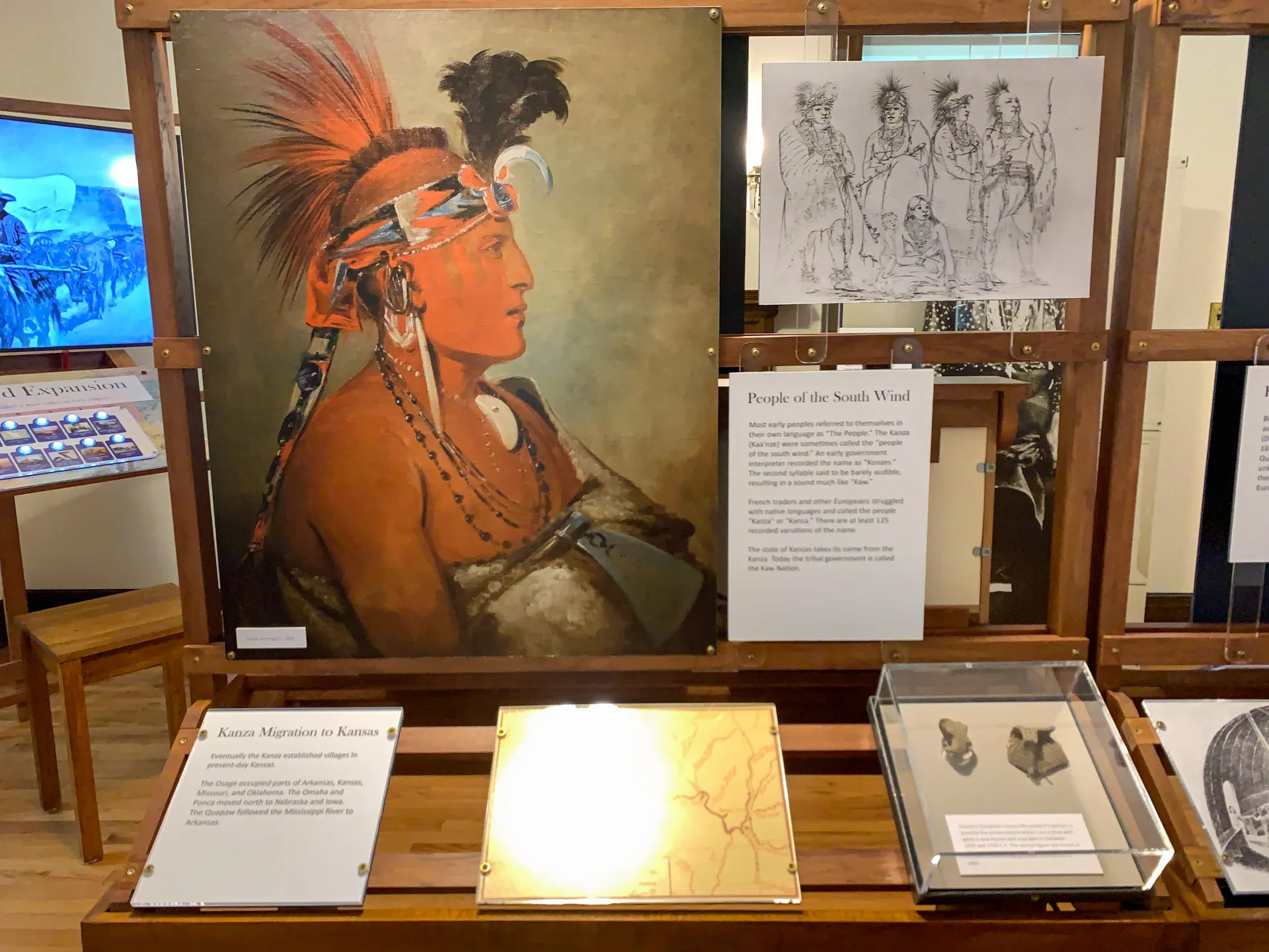 A museum display showcasing a portrait of a Native American man with traditional attire, surrounded by artifacts and descriptions. To the right, a historical sketch and text panel titled "People of the South Wind" are visible. Below are an open book, a map, and small artifacts in glass cases.