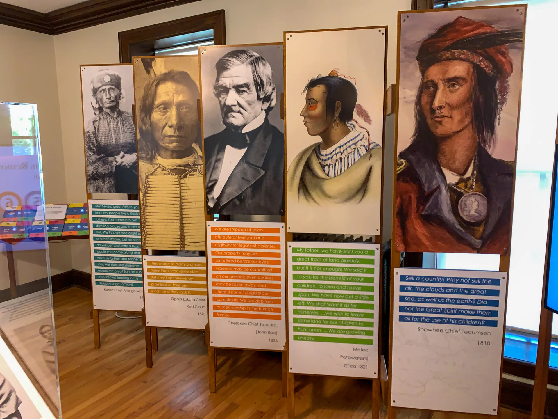 Exhibit showcasing five portraits of Native American and historical figures on standees. Below each portrait, there are colored panels with text about treaties and historical events. The display is set in a room with wooden floors and white walls.