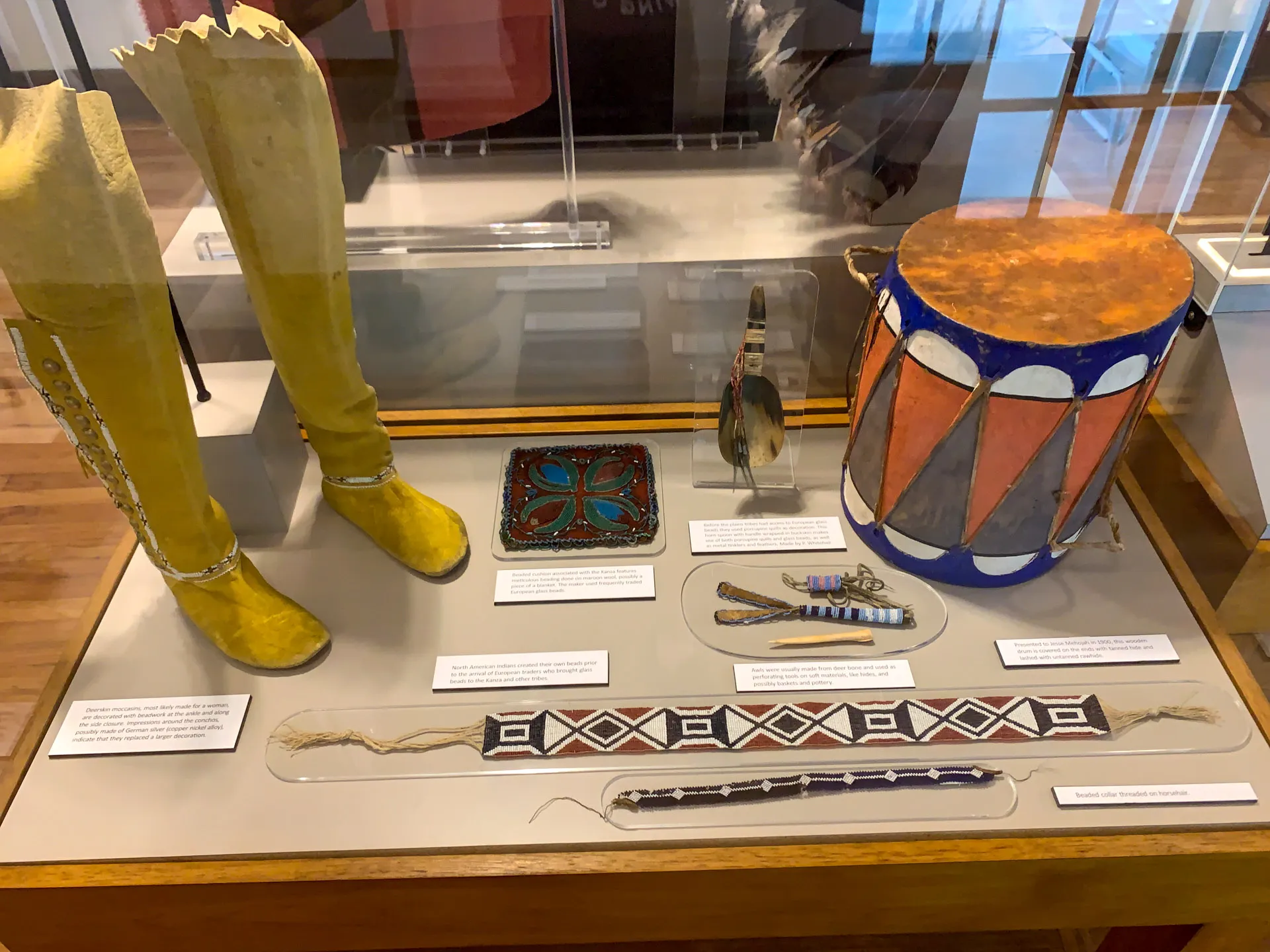 Display case featuring indigenous artifacts: yellow moccasins, a feather, a colorful beaded square, a drum with blue and orange patterns, and intricately beaded belts. Each item has a descriptive label. Wood-framed glass case with a brown floor.