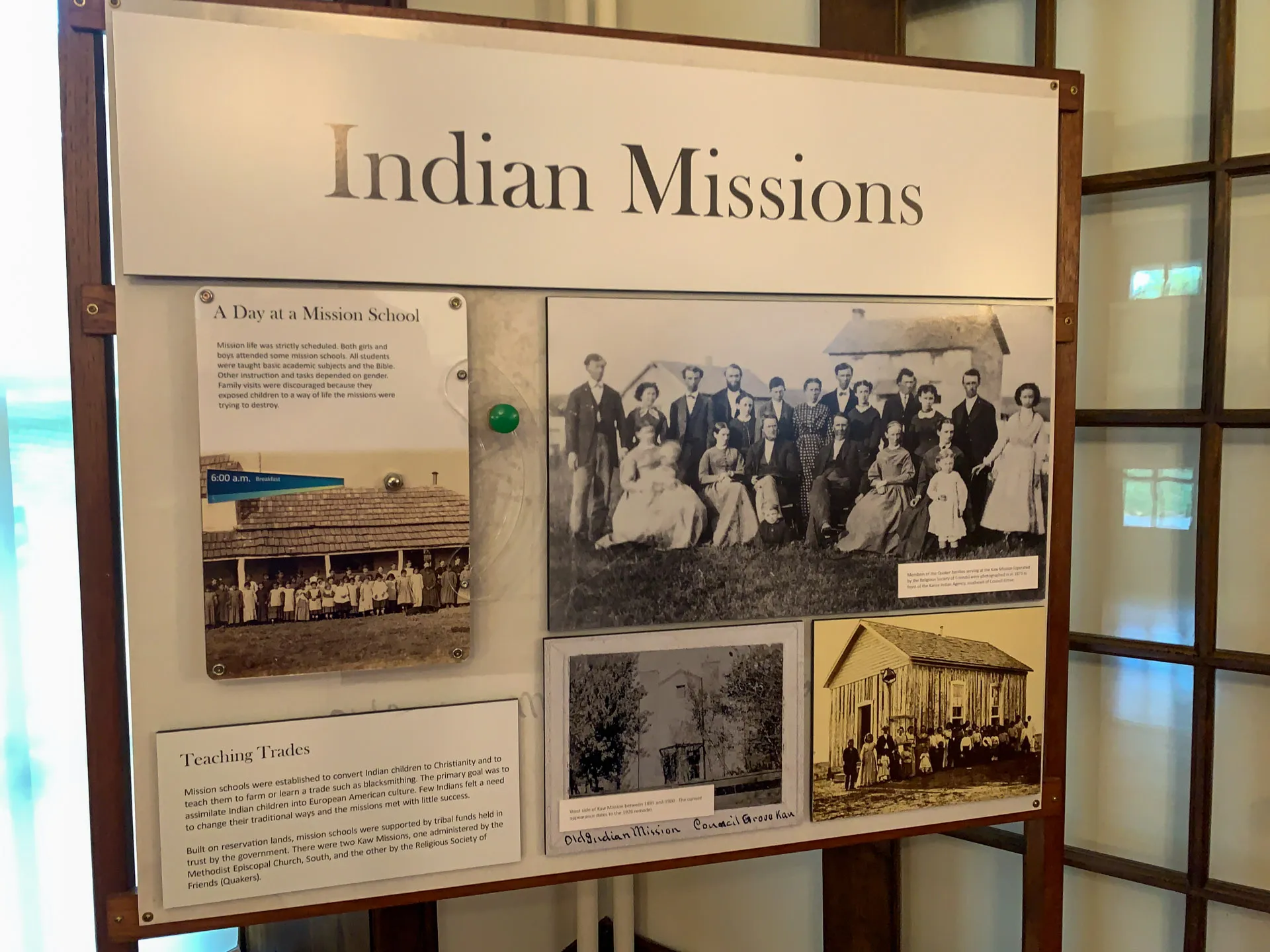 A display board titled "Indian Missions" features historical photos and text. One old photo shows a group of people, another depicts a mission building. Text on the board describes daily life and teaching trades at mission schools. The setting appears to be indoors, in a museum or exhibit space.