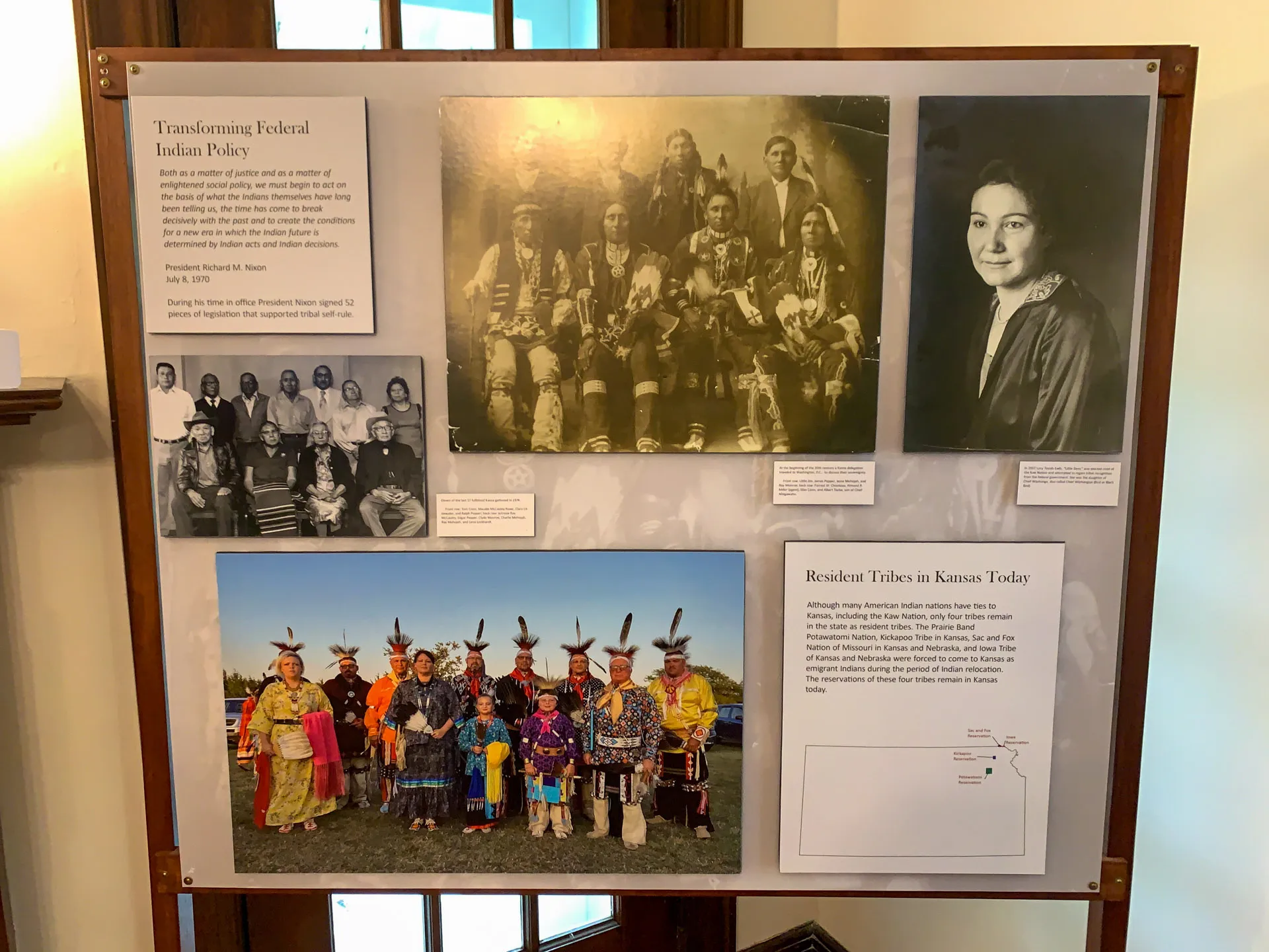 Display board featuring historical and contemporary images of Indigenous people. Top left: federal policy info with a black-and-white group photo. Top right: portrait of a woman. Bottom: colorful photo of tribal members in regalia with text about Kansas tribes today.