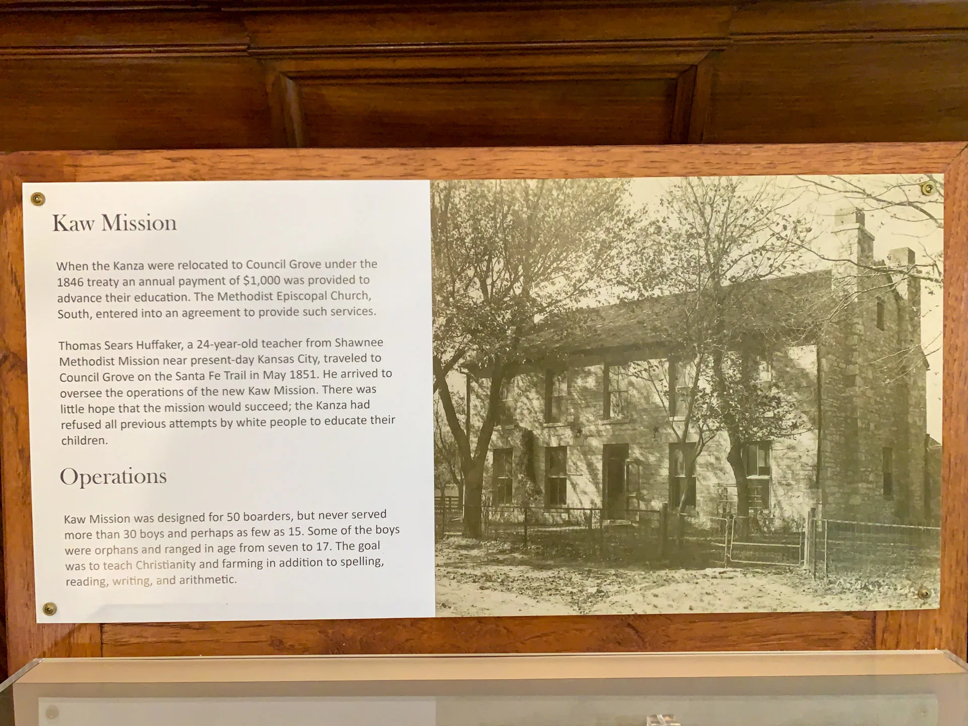 A display with a historical photograph of Kaw Mission, featuring an old building surrounded by trees. The text describes the arrival of Thomas Sears Huffaker in 1851 and the operations of the Kaw Mission, which educated Native American boys. The sign provides details on the mission's history and function.