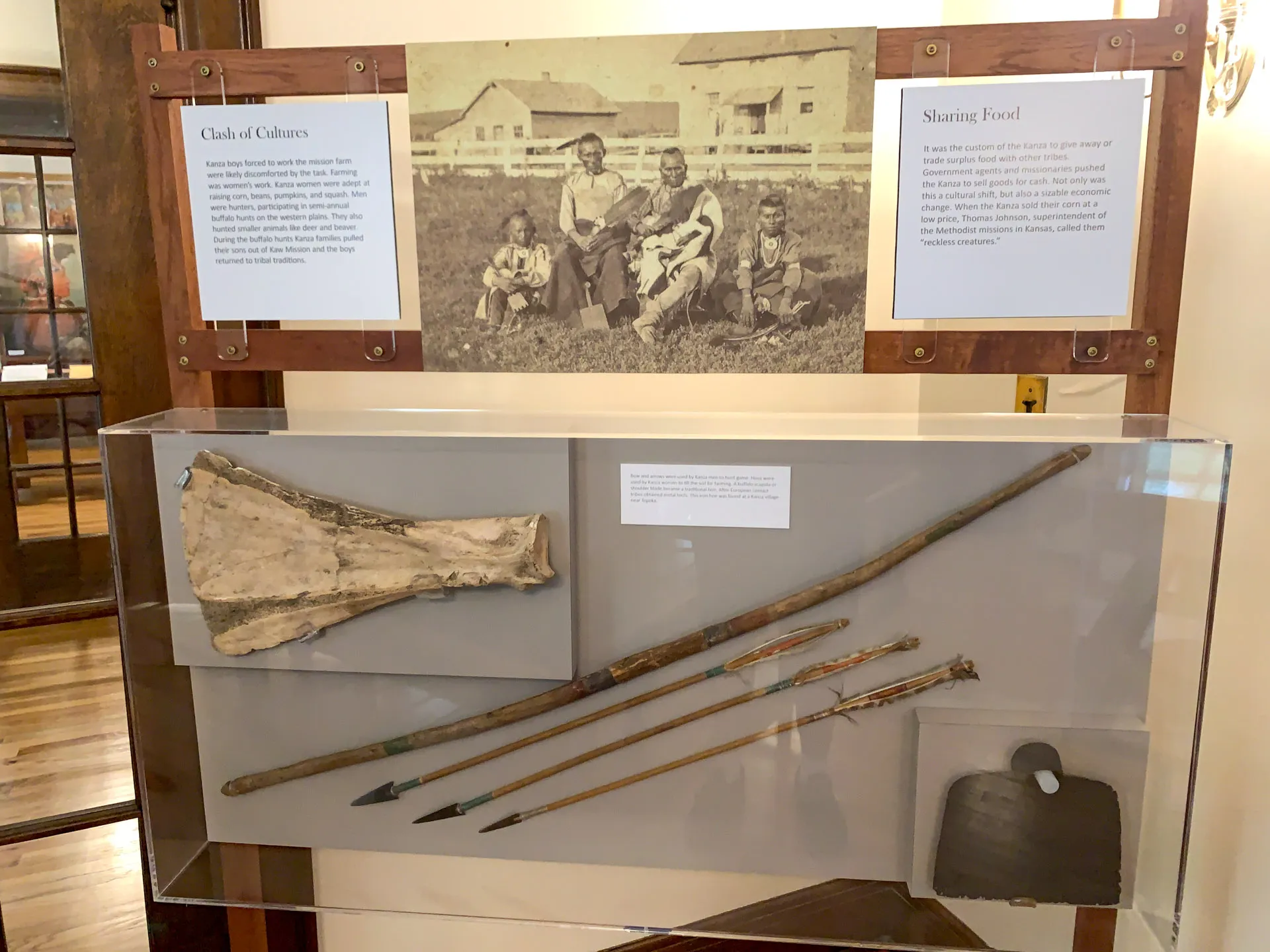 A museum exhibit displays a photo of several people sitting on grass near a wooden fence, titled "Clash of Cultures" and "Sharing Food." Below, a case contains large bones, spears with metal tips, and an iron cooking tool, suggesting historical artifacts of cultural interaction.