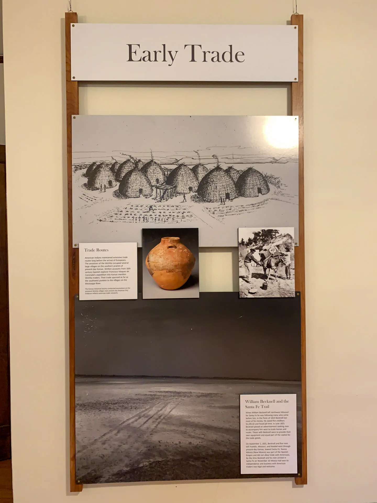 Museum exhibit titled "Early Trade" with images: top section shows drawings of round huts; middle section includes a clay pot and a historical photo of a market scene; bottom section depicts a landscape. Text panels provide context on trade routes and William Becknell’s role in local trade.