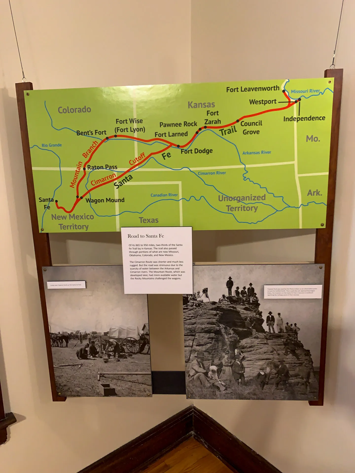 A museum display features a map highlighting the Santa Fe Trail, marking stops from Fort Leavenworth, Kansas, to Santa Fe, New Mexico, with a red line. Below the map are two historical photos: one of wagons and settlers, and another of people gathered on a rocky terrain.