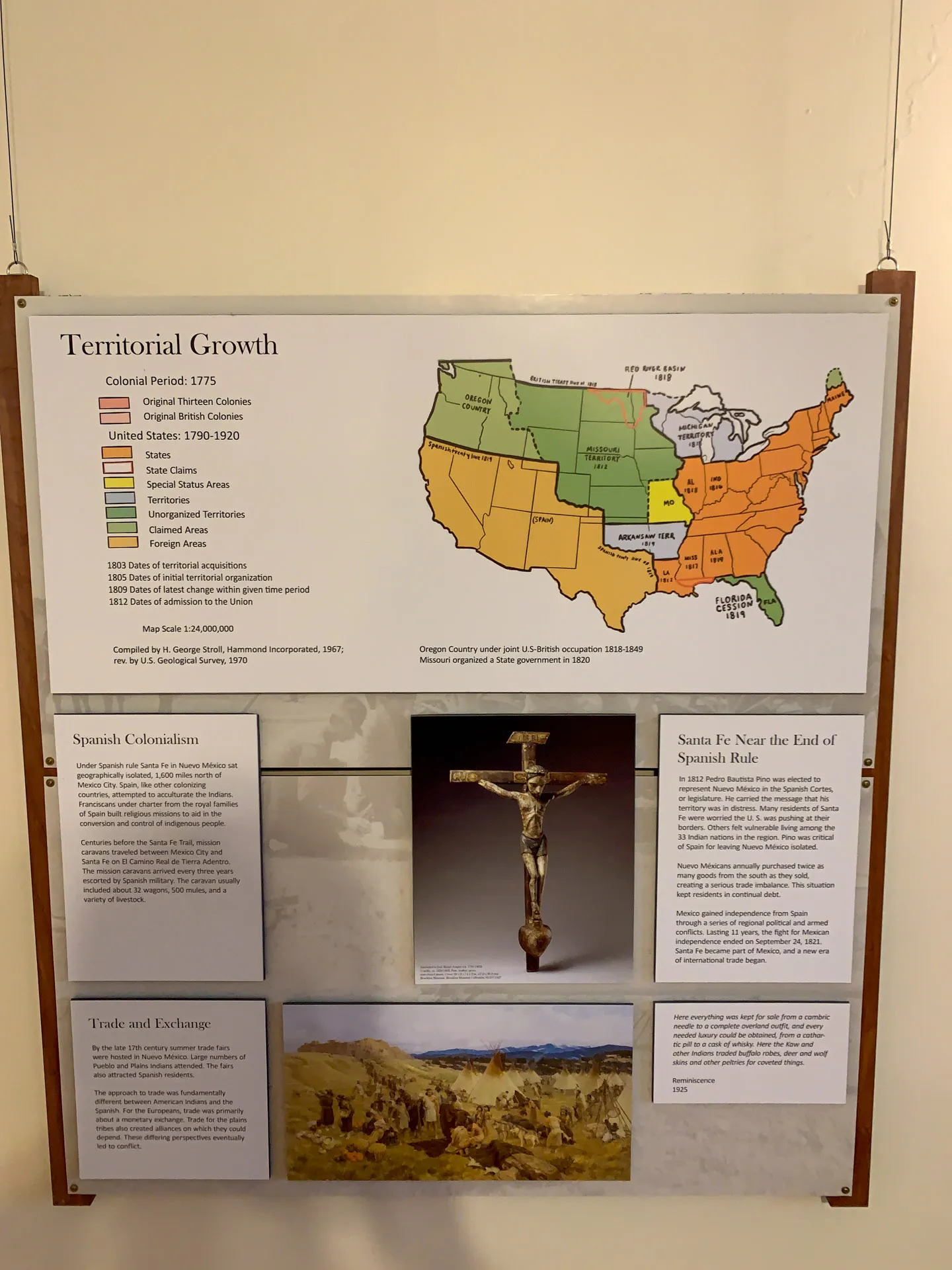 A historical display board shows a U.S. map illustrating territorial growth, color-coded by periods. Below are sections titled "Spanish Colonization," featuring text and an image of a cross, and "Trade and Exchange," with a desert landscape photo. The board hangs on a beige wall.