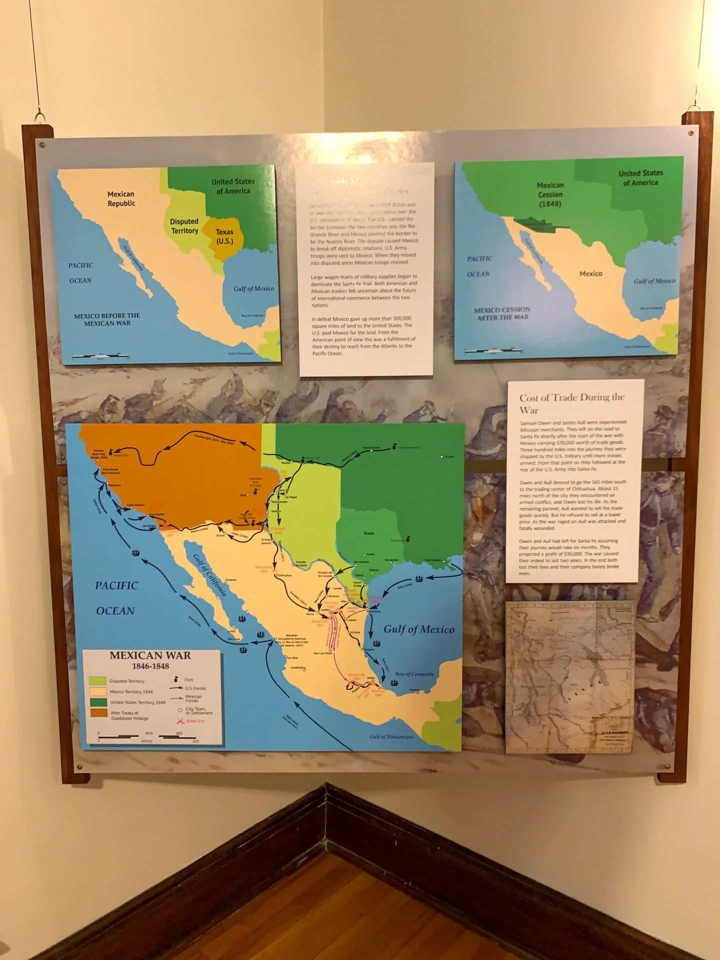 Exhibit of maps and text detailing the Mexican-American War. Includes colorful maps showing territorial changes and historical routes, a text box on trade costs, and descriptions. Wooden floor and cream-colored walls frame the display.