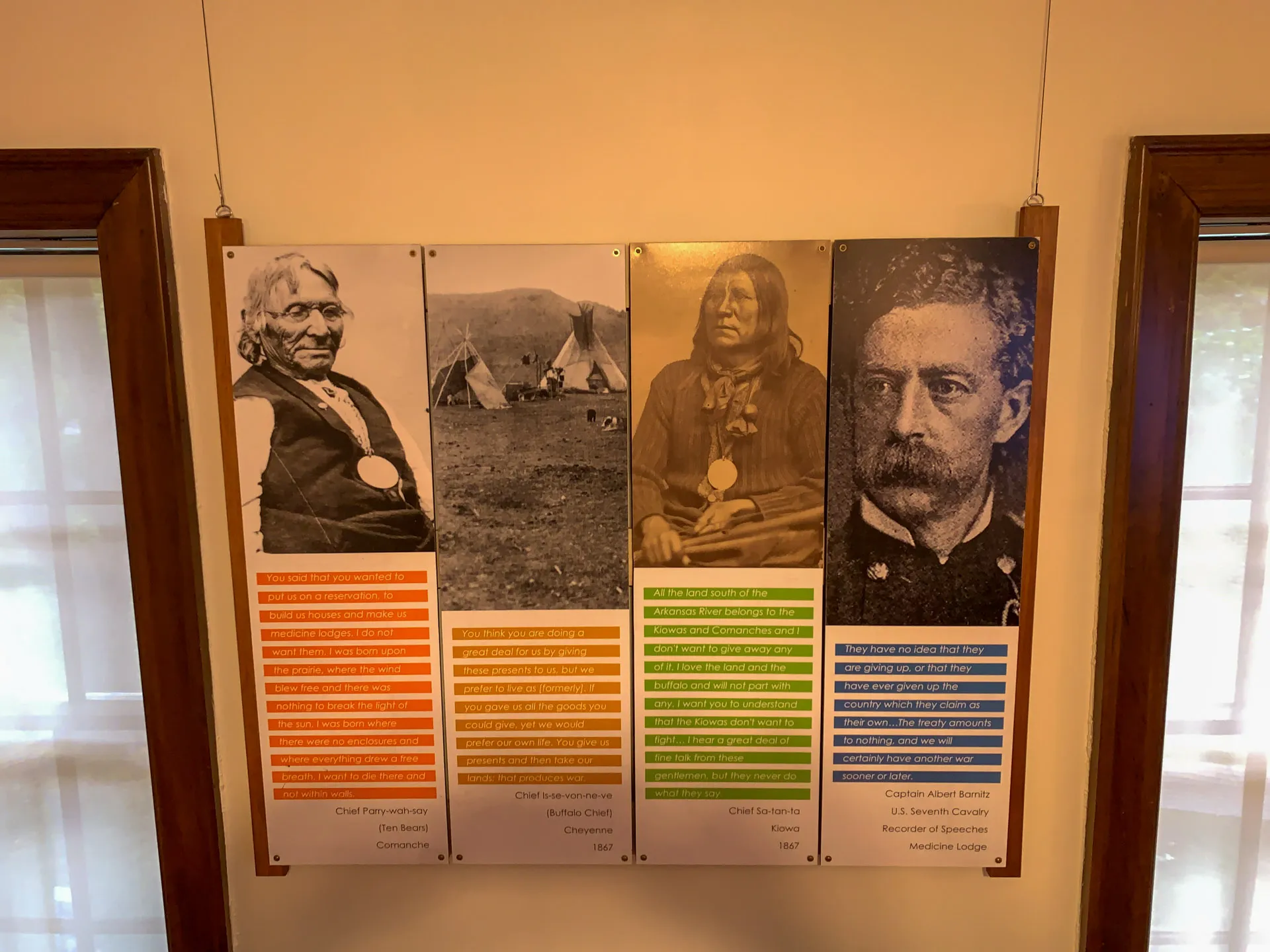 A museum display features four vertical panels with images and text. From left to right: portraits of Chief Plenty Coups, Buffalo Calf Road Woman, Chief Sam Sinquesip, and Captain Robert Snyder. Below each image is a biography and achievements highlighted in various colors.