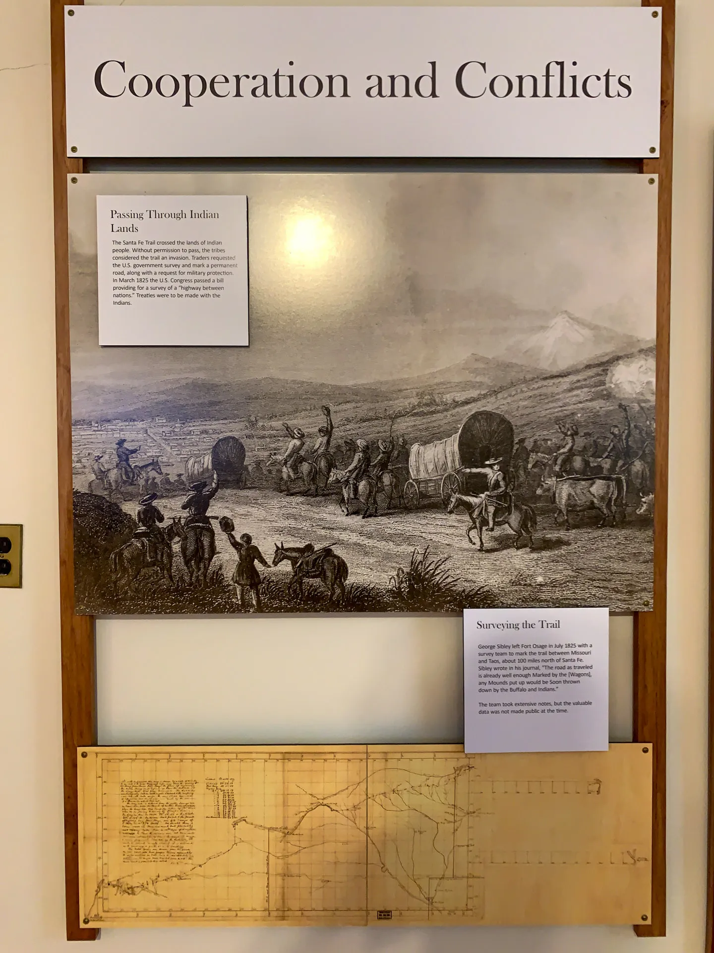 A museum display titled "Cooperation and Conflicts" features two framed sections. The top section shows an illustration of settlers with covered wagons crossing a landscape, with Native Americans observing. The bottom section contains a historical map. Text descriptions accompany both sections.
