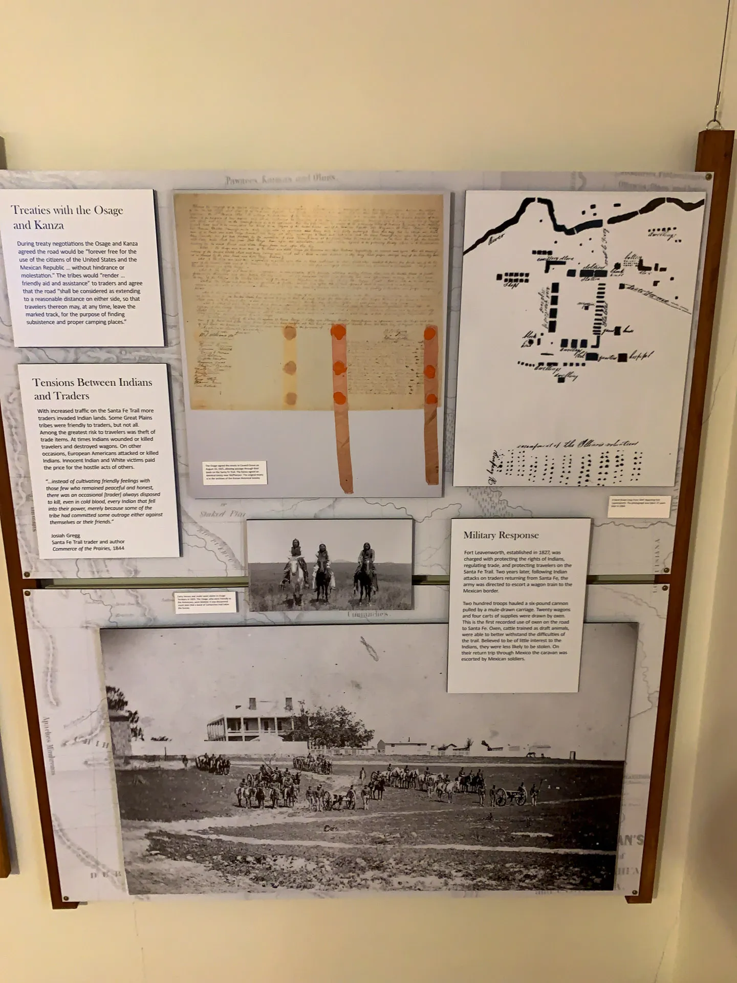 A museum display showcases historical documents and images. It includes a hand-drawn map, text panels titled "Treaties with the Osage and Kansa" and "Tensions Between Indians and Troops," a photo of Native American riders, and an image depicting a military fort with soldiers and horses.