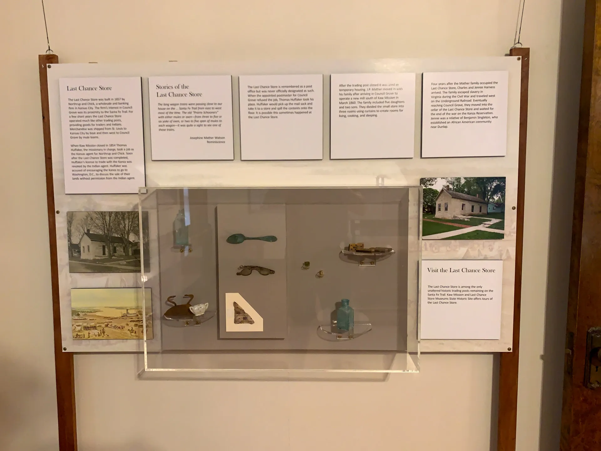 A museum display features a wooden frame with six informational panels about the Last Chance Store. Below, a clear case contains historical artifacts, including utensils, a blue glass bottle, and assorted items. An image of the store is displayed in the bottom right panel.