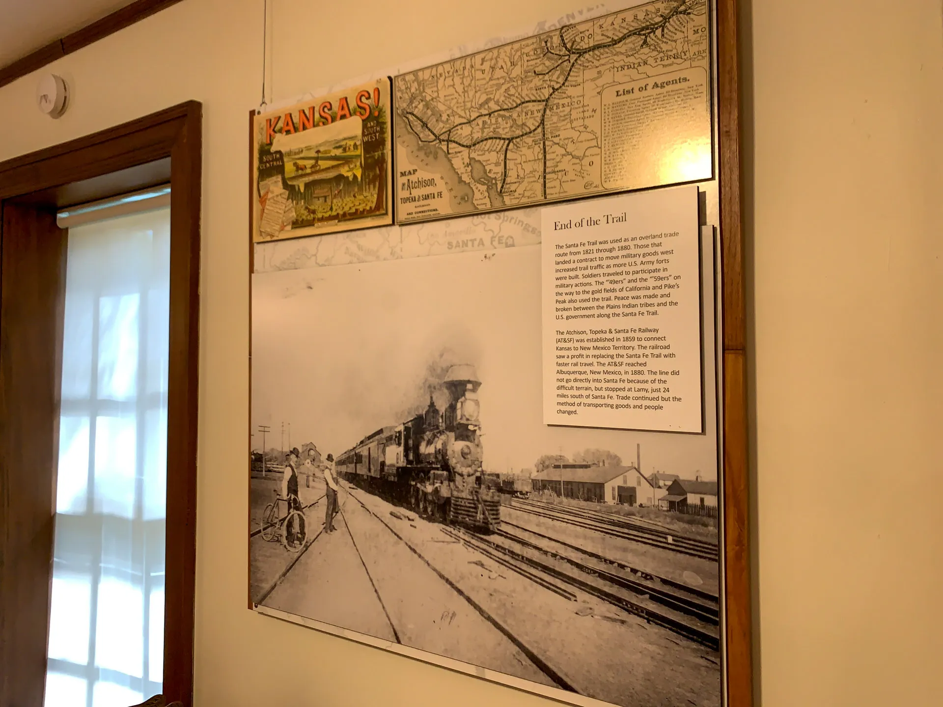 Wall display featuring a historic black-and-white photo of a steam locomotive at a station. Above it are map sections, a vintage Kansas postcard, and a text panel titled "End of the Trail" describing the railroad's history. The display is in a warmly lit, wooden room.
