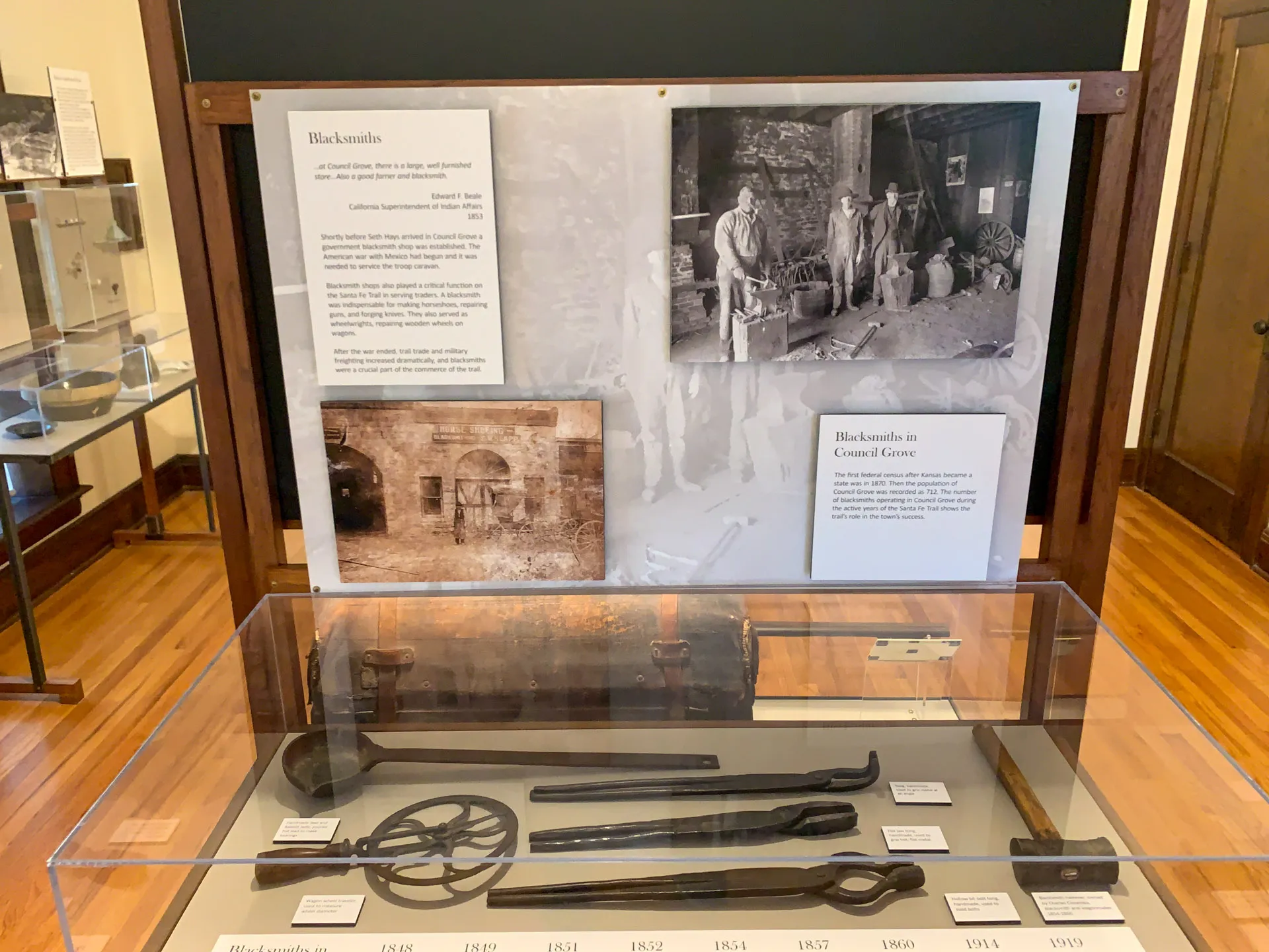 A museum exhibit displays blacksmithing tools, including tongs, a circular tool, and a long wrench, encased in glass. Above are informational panels and historical black-and-white photos of blacksmiths at work. The exhibit is on a polished wooden floor.